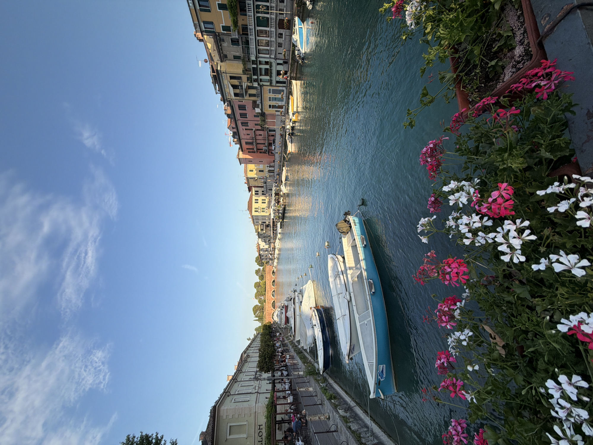 The image depicts a scenic view of a canal in Peschiera del Garda, Italy, likely during the late afternoon or early evening based on the lighting. The canal is lined on the left by buildings with awnings and outdoor seating areas where people are visible. Several boats are moored along the canal, including a prominent blue boat in the foreground. The water is calm and reflects the sky and the colorful buildings on the right side of the canal. These buildings are varied in height and color, featuring balconies and windows. In the distance, a stone bridge with multiple arches spans the canal, and beyond that, more buildings and trees are visible. The sky is a clear blue with a few wispy clouds. In the immediate foreground, a planter box filled with blooming pink and white flowers adds a touch of color and softness to the scene. The overall atmosphere is peaceful and picturesque.