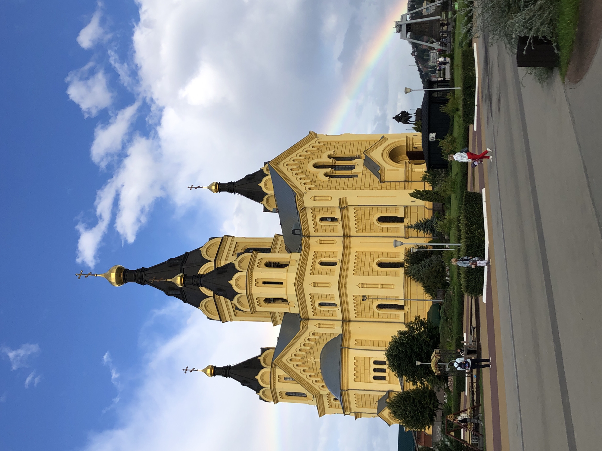 The image shows the Alexander Nevsky Cathedral in Nizhny Novgorod, Russia, a prominent landmark in the city. The cathedral is a large, yellow Orthodox church with multiple black domes topped with golden crosses. A rainbow is visible in the sky to the right of the cathedral, arching over the landscape. In front of the cathedral, there is a park-like area with trees, bushes, benches, and lampposts. Several people are present in the park: a person wearing a backpack walks on a paved path, another person with a light-colored jacket walks towards the right, and a person in a white jacket and red pants is walking with a bouquet of flowers in their left hand. Further in the background, a statue of a man on horseback is visible, and beyond that, a large bell structure can be seen. The sky is partly cloudy with patches of blue. The foreground consists of a paved area, likely a pedestrian walkway.
