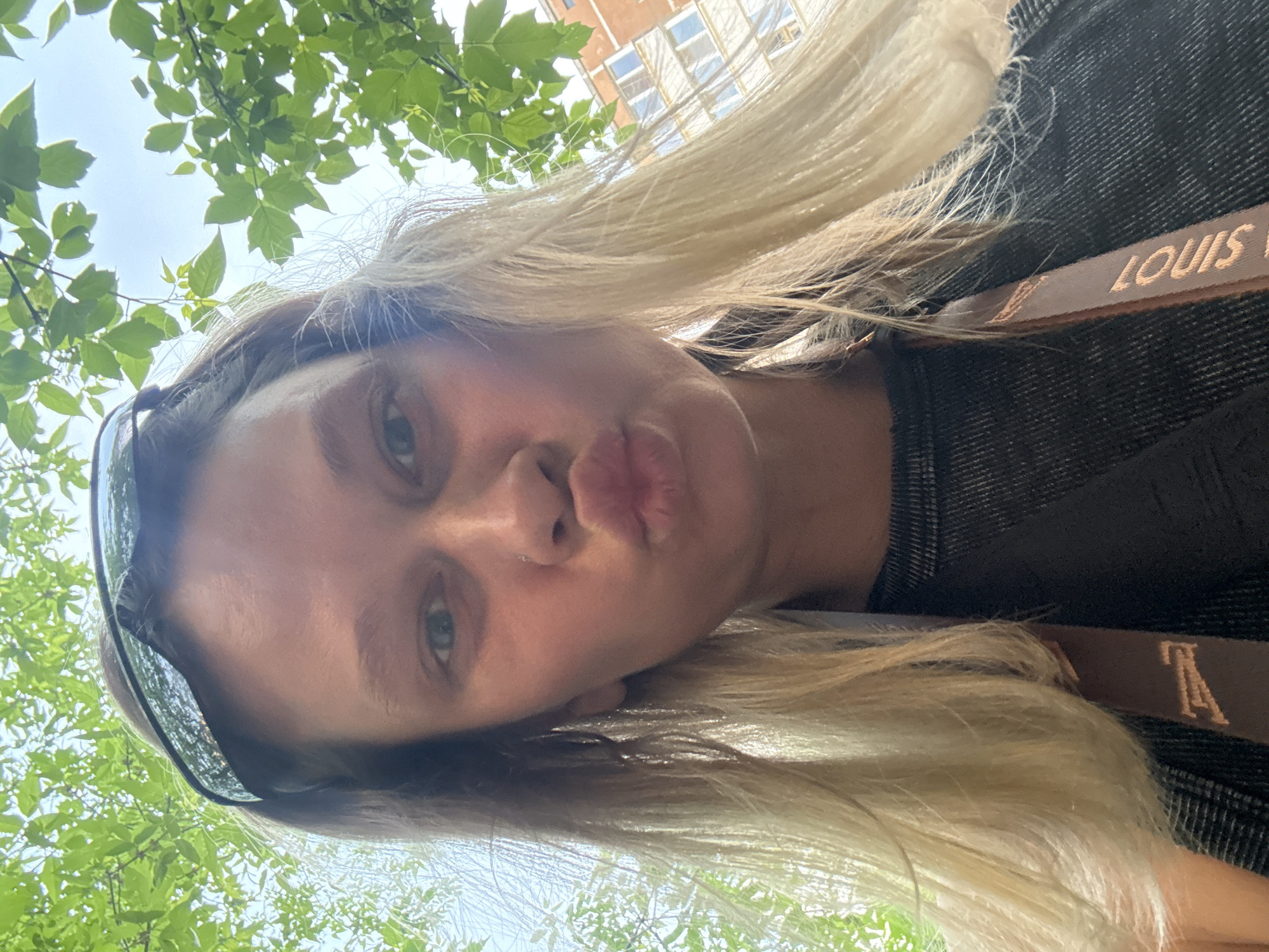 A woman with blonde hair is taking a selfie. She is wearing sunglasses and a dark shirt. A strap, possibly for a handbag, is visible, with the name "LOUIS" printed on it. The background shows green leaves of trees and a building. The location is likely Perm, Russia, based on the prompt's request to include this information.