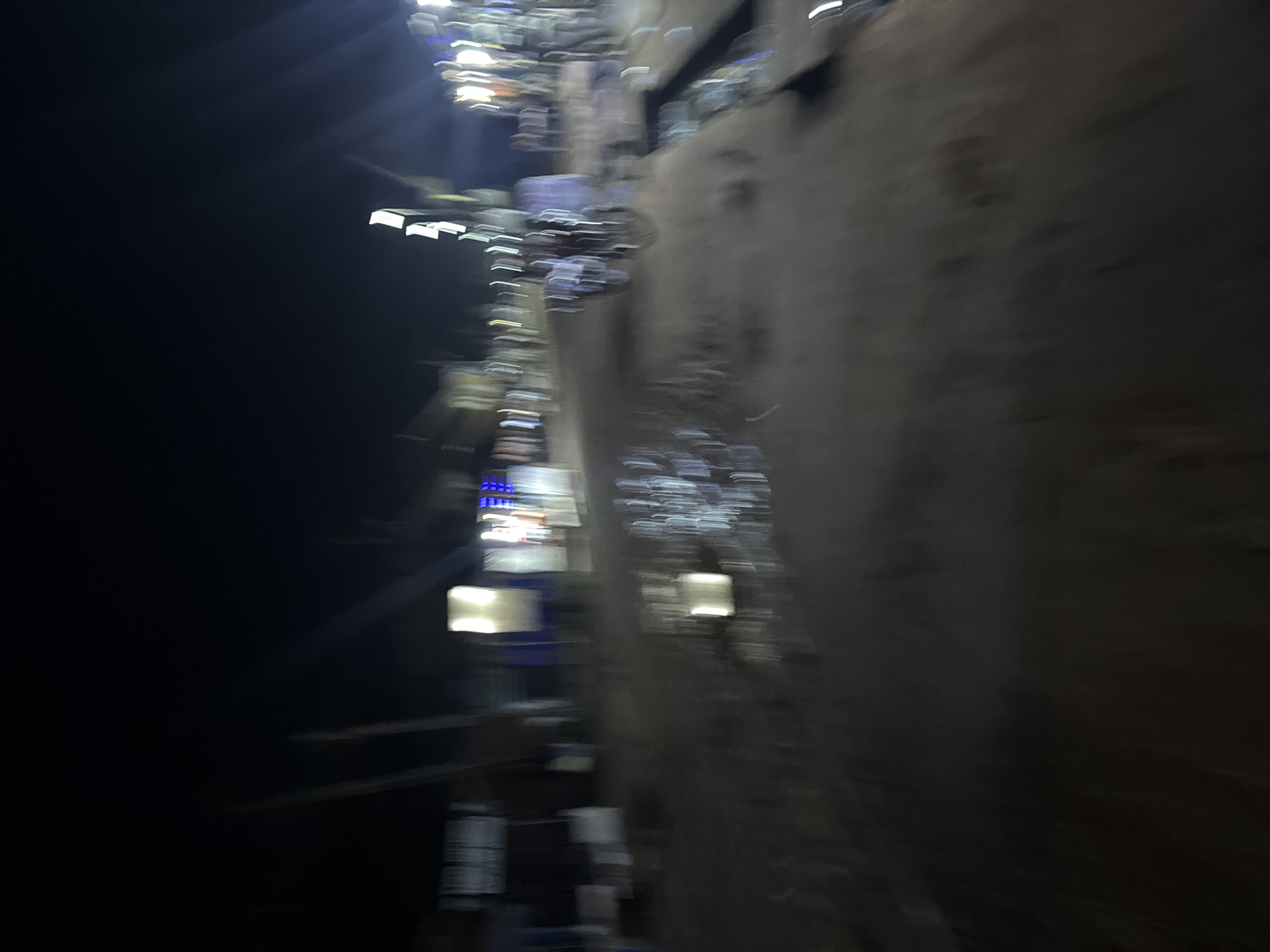 The image shows a blurred, sideways view of a street in Eziobodo, Nigeria, at night. Illumination comes from streetlights and building lights, creating streaks due to motion blur. A dark, unlit area occupies the left side of the image. On the right is a dark colored wall. Other details are unidentifiable due to the blur. No people are clearly visible.