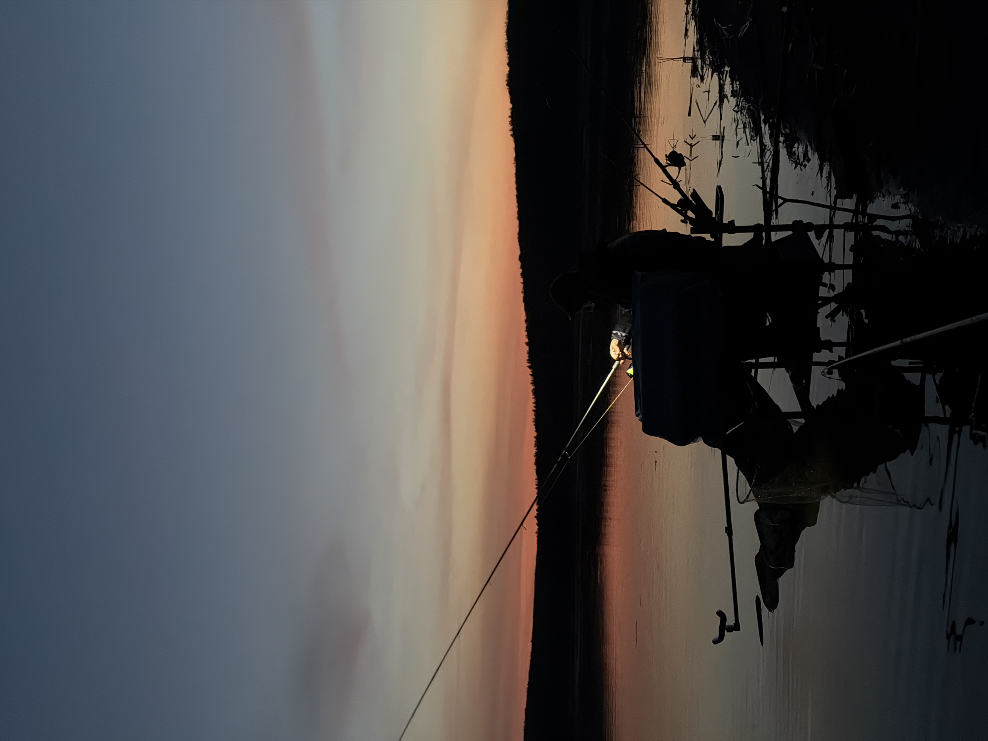 The image depicts a silhouetted figure fishing at dusk in Chaykovsky, Russia. The sky transitions from a deep blue on the left to a warm orange and pink on the right, possibly indicating sunset or sunrise. A dark line of trees or land separates the sky from the water. In the foreground, multiple fishing rods are visible, some with glowing tips or lights attached. The fisherman appears to be seated or standing near the water's edge, with various fishing equipment and possibly a tackle box nearby. The silhouette suggests a focus on the activity of fishing against the backdrop of the natural environment. The overall scene captures a sense of serenity and solitude.