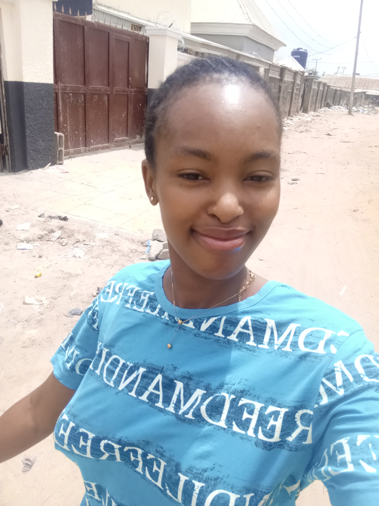 A young woman is taking a selfie on a dirt street in Keffi, Nigeria. She has dark hair pulled back from her face, and is wearing a light blue t-shirt with white lettering. She is wearing a gold necklace. The background shows a brown metal gate, a concrete wall topped with barbed wire, utility poles, and low buildings. Debris litters the street.
