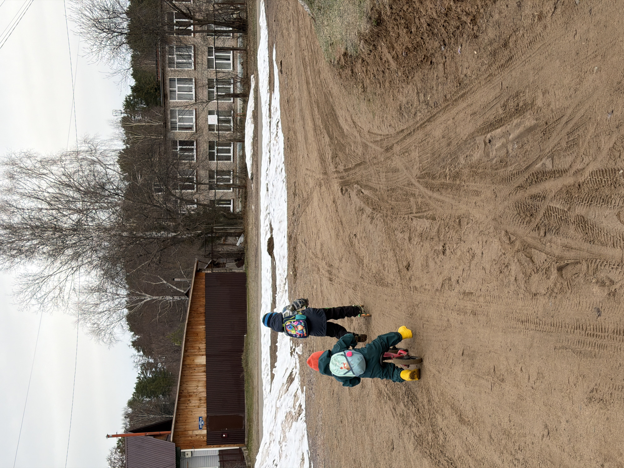 The scene is a dirt road in the city of Chaikovsky, Russia. Two young children, wearing warm clothing and backpacks, are walking on the road. One child is on a balance bike. The road is sandy with many tire tracks. Patches of melting snow are visible along the side of the road next to several buildings. A multi-story brick building stands to the left of the frame and a wood plank structure with a brown roof, presumably a house, is visible at the very bottom left. The sky is overcast. Bare trees surround the buildings, indicating it is early spring or late fall.