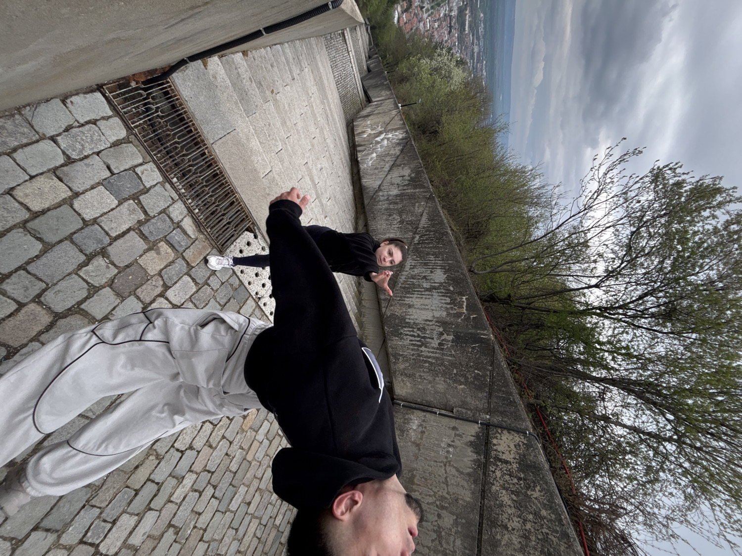 This image is a photograph taken from a low perspective and rotated 90 degrees clockwise, depicting two individuals on an outdoor paved path overlooking a distant cityscape.
In the foreground, an individual is captured in an inverted position, suggesting they are performing a handstand or a similar acrobatic pose. They are wearing a black long-sleeved top, possibly a hoodie, and light grey or white trousers with distinctive black outlining or seams. Their head is partially visible near the bottom edge of the frame. The ground beneath them consists of grey cobblestones and a section of concrete. To their right, a metal grate, likely a storm drain, is embedded in the pavement.
Further along the path, towards the mid-ground, a second individual is visible, standing upright relative to the path but appearing sideways due to the image rotation. They are looking directly at the camera with a slight smile and extending their left hand in a gesture, with fingers spread. This person is wearing a dark-colored top and white sneakers are visible on the concrete section of the path near their feet.
The setting is outdoors, suggesting a viewpoint or a path winding up a hillside. A concrete wall runs along the right side of the path, with trees and sparse vegetation growing beyond it, indicating a natural landscape. In the far background, a sprawling city or town is discernible beneath a cloudy, overcast sky, suggesting it is daytime. The scene conveys a casual and playful atmosphere, capturing an interactive moment between the two individuals in a scenic, elevated location. No visible text is present in the image.