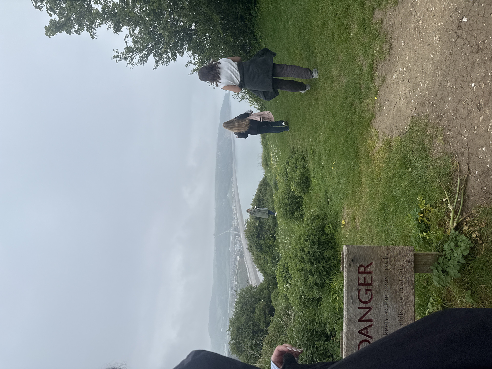 The image shows a coastal scene in East Devon, United Kingdom, taken from a high vantage point. Two females stand near the edge of a grassy cliff, overlooking a town along the coastline. Below them, another person stands on the slope. A wooden sign with the word "DANGER" is visible in the foreground, warning people to keep to the coast path because the cliffs are unstable. The day appears overcast.