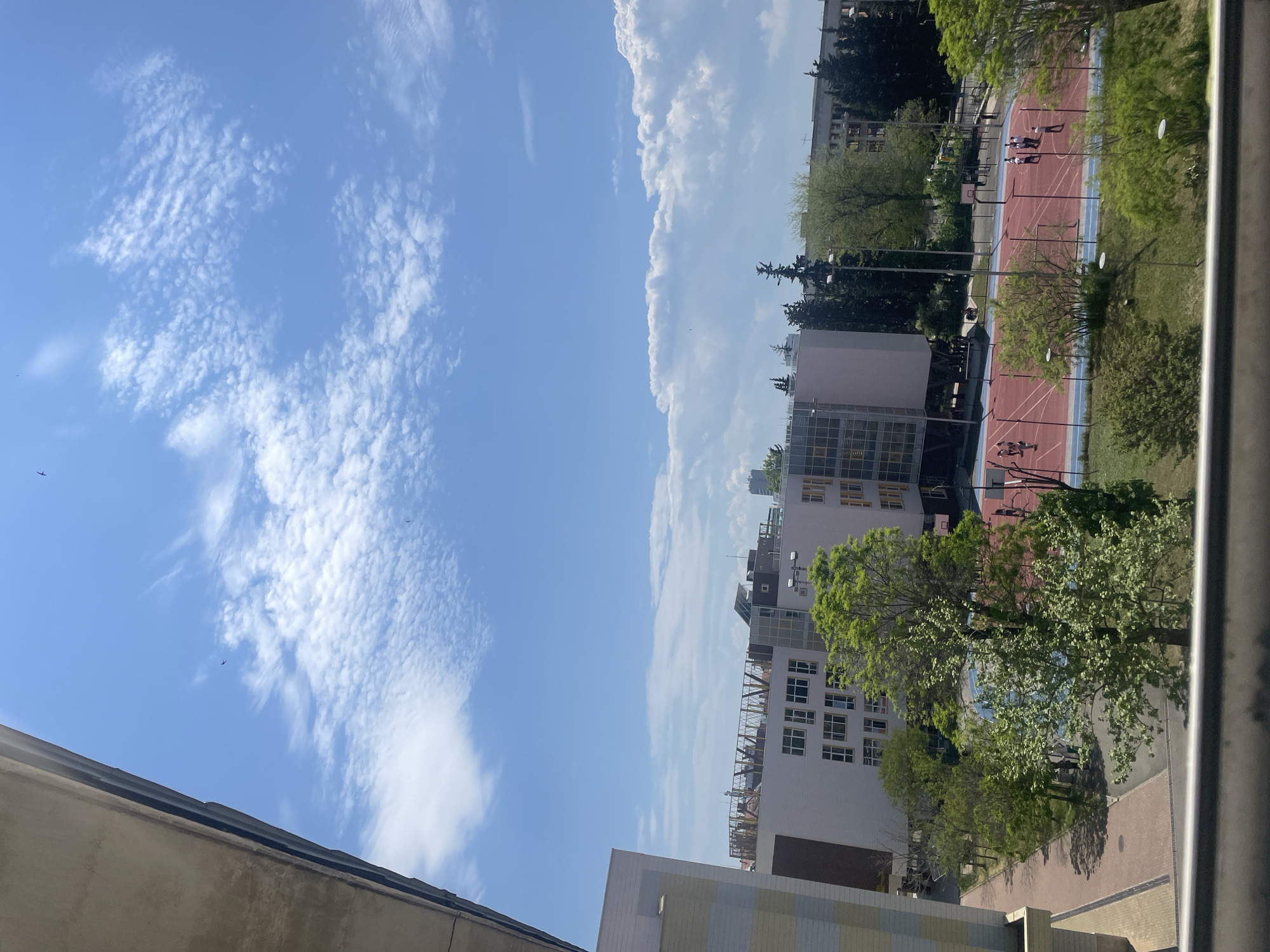 The image presents a partial view of an urban environment under a blue sky with scattered clouds. Buildings of varying heights and architectural styles are visible, including a multi-story structure with numerous windows. A running track, marked in red and blue, is adjacent to the buildings and features several individuals engaging in sports activities. Trees with green foliage are interspersed throughout the scene, adding a natural element to the urban landscape. A section of a roof and a railing are visible in the foreground, suggesting the vantage point is from an elevated position, possibly a balcony or upper floor of a building.