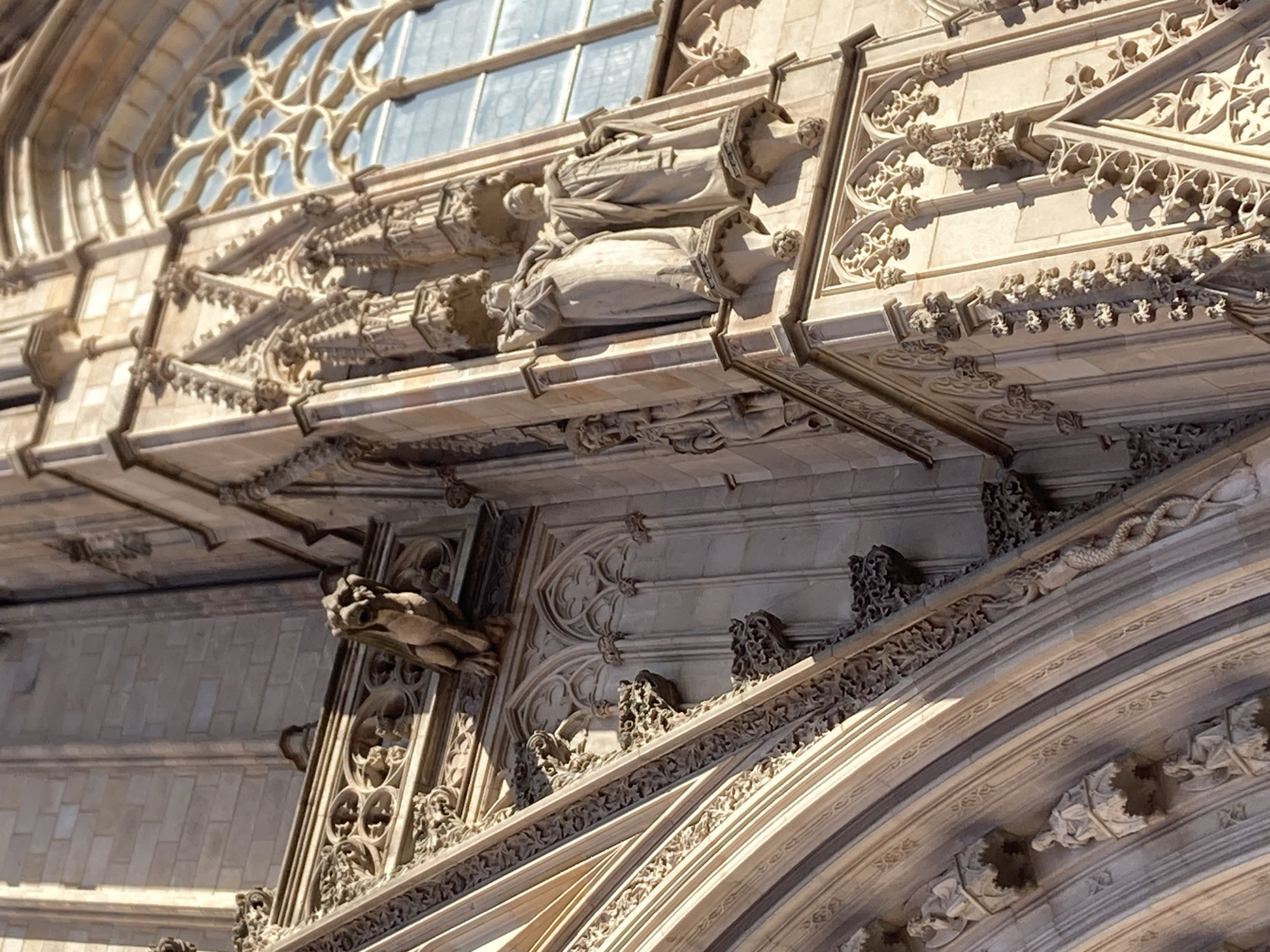 This media file features the exterior architectural details of a gothic building, likely a cathedral, in Barcelona, Spain. The scene focuses on intricate stonework, specifically carved figures and decorative elements adorning the facade. Prominently visible are two statues of robed figures, one above the other, resting on sculpted bases. Below these figures, a grotesque gargoyle-like carving emerges from the stonework, its features weathered and detailed.
The architecture exhibits characteristic gothic elements such as pointed arches, tracery in the windows (partially visible at the top of the frame), and ornate sculptural embellishments along cornices and ledges. Small, decorative carvings of foliage and mythical creatures, including a serpent-like figure, are interspersed throughout the facade. The stonework appears to be made of light-colored sandstone, with some areas showing signs of age and wear.
The lighting suggests it is daytime, with strong sunlight casting distinct shadows that highlight the three-dimensional nature of the carvings and the depth of the architectural features. The clear illumination indicates fair weather. The overall impression is one of grandeur and historical significance, characteristic of major European religious or civic buildings. There is no visible text in the image.