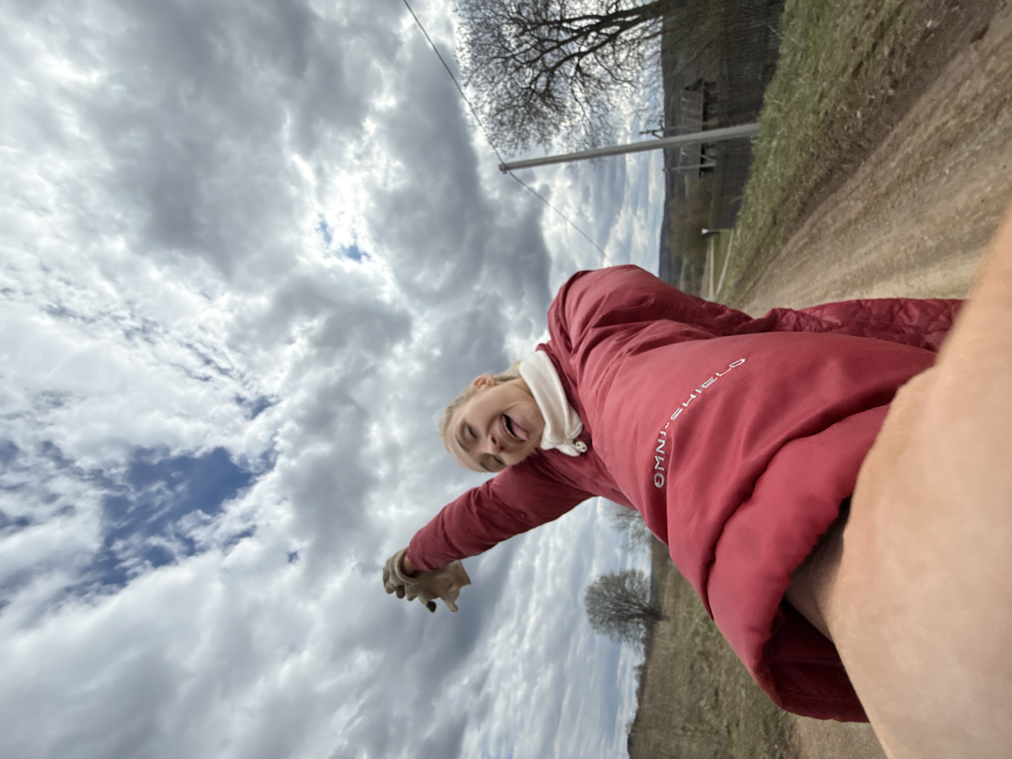 A young woman is captured in a playful selfie, taken from a low, upward-looking angle and rotated 90 degrees clockwise, making the horizon appear vertical. She is smiling broadly and sticking her tongue out, conveying a cheerful and energetic mood. Her blonde hair is pulled back, and she wears a light-colored hooded top under a red puffer-style jacket. The text "OMNI-SHIELD" is visible on the right sleeve of her jacket. She has a gloved left hand reaching out towards the camera, while her right arm (carrying the camera/phone) is mostly obscured.
The setting is outdoors in what appears to be a rural or semi-rural area in Rosstani, Russia. The background features a dirt path or road to the right, bordered by some grassy and bare ground. In the distance, hills or mountains are visible under a vast, cloudy sky with patches of blue. Several bare-branched trees and a utility pole are also part of the landscape. The weather is overcast but bright, suggesting daytime.