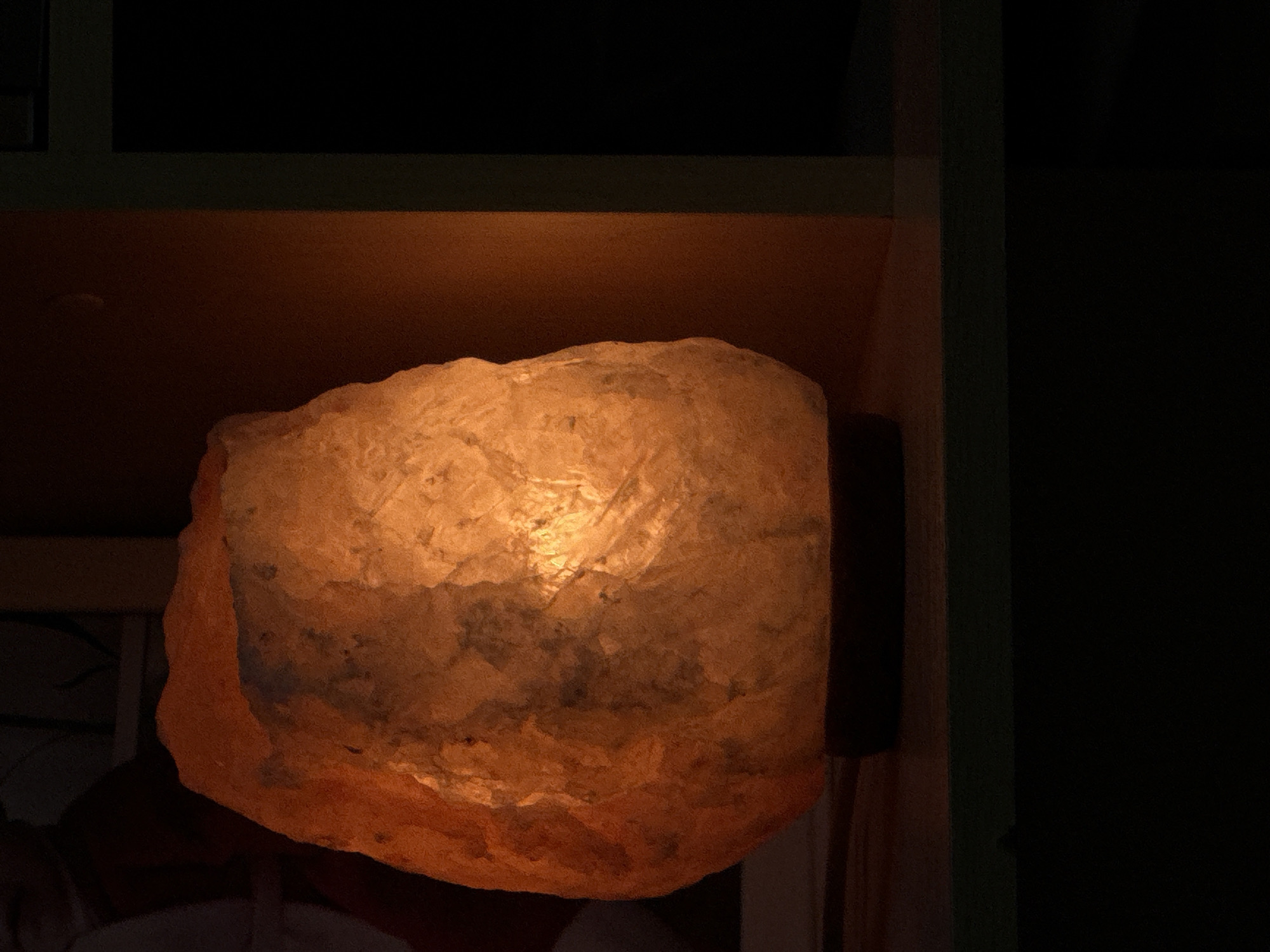Illuminated salt lamp within a dimly lit interior, positioned near a wall and a green frame. The surroundings are dark.