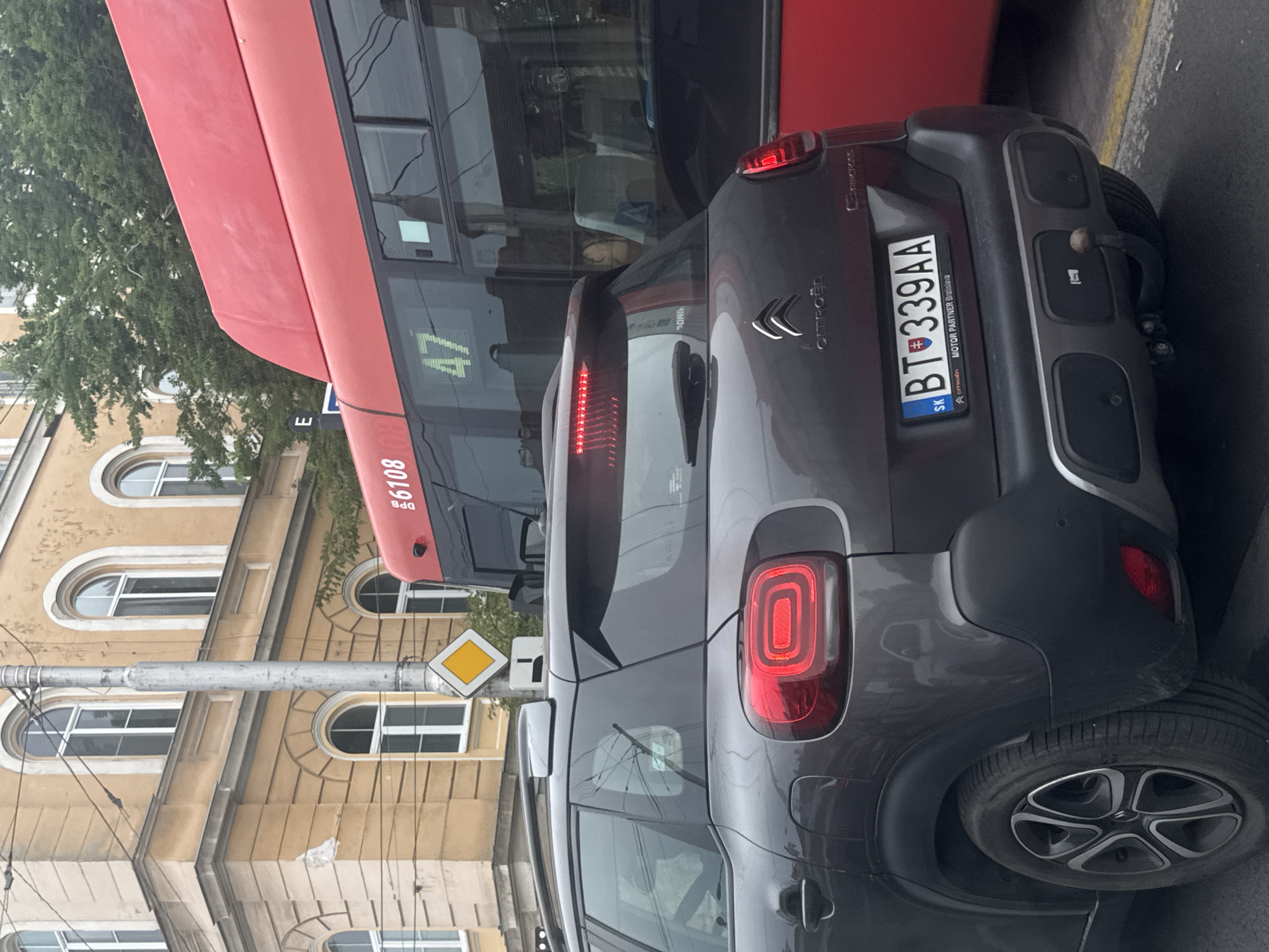 The image shows a dark gray Citroën C3 Aircross parked on a street, likely in Bratislava, Slovakia, based on the license plate "BT 339AA" which is a Slovakian format. The car is viewed from the rear and is positioned to the left of a bright red bus. The bus has the number "47" displayed prominently on its side, and what appears to be a route number or identifier "6108" on its front.
The background features a yellow building with arched windows and architectural details typical of older European city structures. A metal pole with traffic signs, including a yellow diamond shape, stands near the car. Trees are visible behind the building and the bus. The street surface is asphalt, with yellow lines marking a lane. The overall lighting suggests an overcast day. The car's rear lights are illuminated, indicating it might be stopped or braking. The focus is on the rear of the car and the bus, with the building and surrounding elements providing context. There are no people clearly visible in the image. The scene appears to be a moment captured in urban traffic.