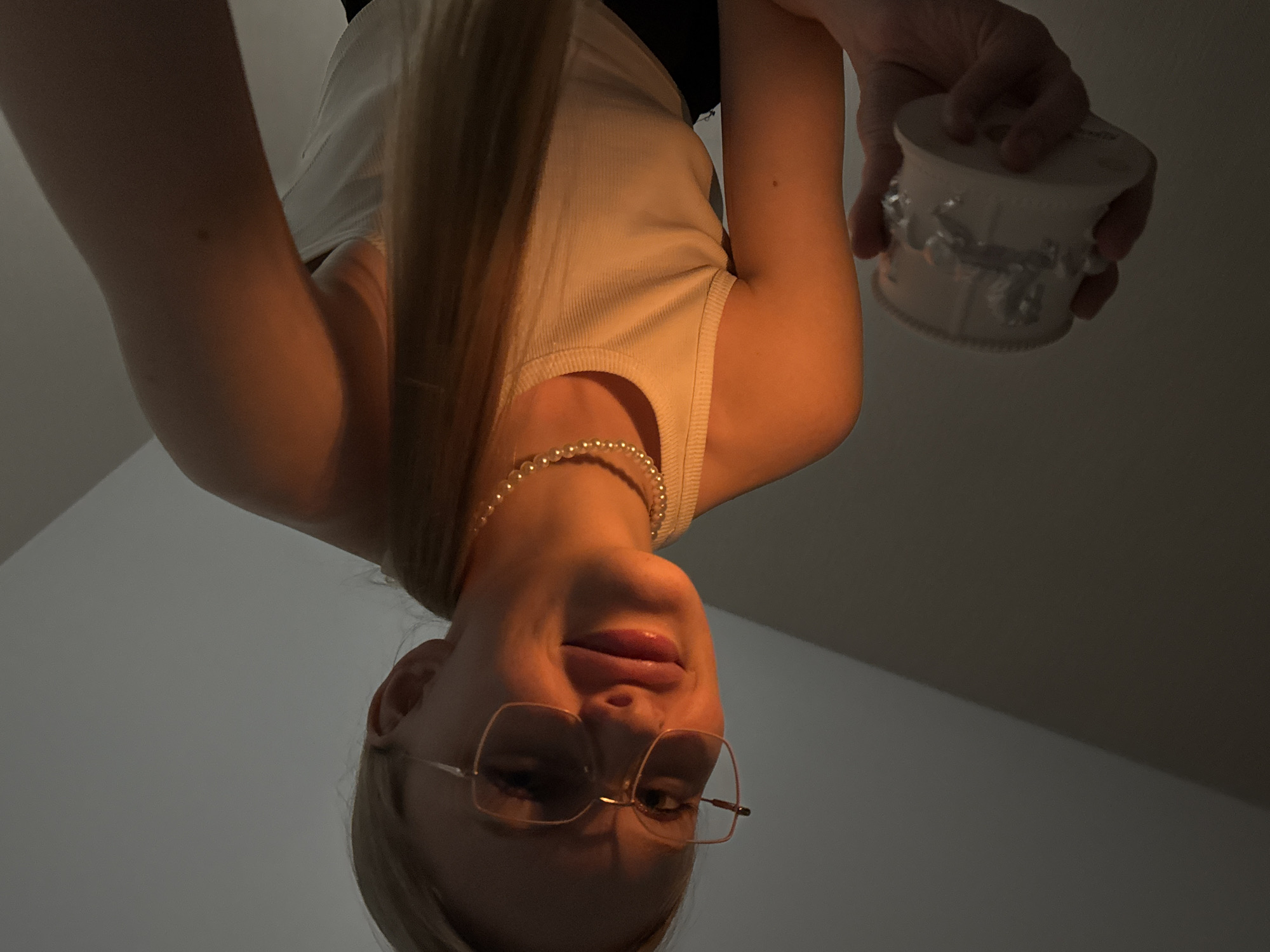 The image, presented upside down, captures a young woman in an indoor setting from a low-angle perspective. If rotated to a standard orientation, she is shown from the chest up, looking slightly downwards with a gentle smirk. She has long, light-colored hair and wears a light-colored, ribbed tank top, a pearl necklace, and rectangular-framed glasses.
She holds a small, ornate, light-colored circular container, possibly a decorative box, in her right hand. A warm, orange-toned light illuminates the left side of her face and arm, creating a strong contrast with the dimly lit or shadowed right side. The background is a plain, light gray surface, suggesting a simple, uncluttered interior. There are no specific visual cues to indicate the stated location of Perm, Russia. No text is visible in the image.