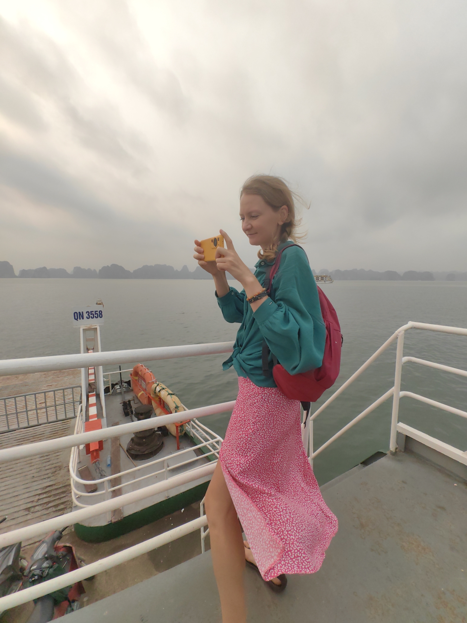 On a cloudy day in Tuan Chau Ward, Vietnam, a woman stands on a ferry and uses a smartphone to take a photo of the scenic view. She has fair skin and light-colored hair, styled in braids, and is wearing a blue-green shirt tied at the waist, a long, pink printed skirt, and a red backpack. The ferry has white railings and a white and green hull, with the number "QN 3558" displayed on a sign. Life preservers are stacked on the ferry deck. The background shows a body of water with several islands in the distance.