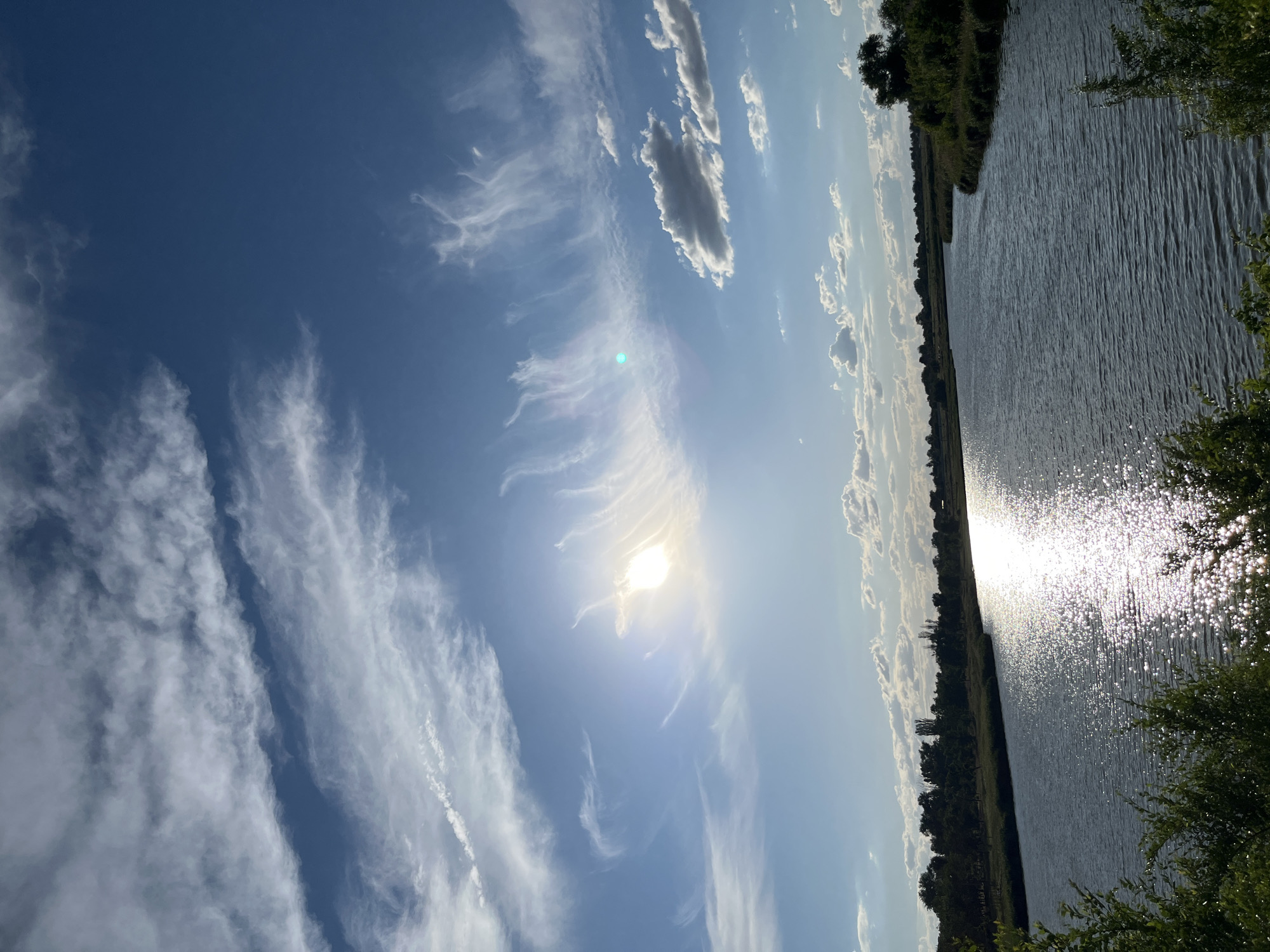 The image shows a wide, calm river flowing through a landscape with trees and vegetation along its banks. The sun is shining brightly, creating a strong reflection on the water's surface. The sky is a clear blue with scattered, wispy white clouds. The scene appears to be a natural outdoor setting, possibly a park or a rural area. The crops suggest the location is Shyroka Dacha, Ukraine.