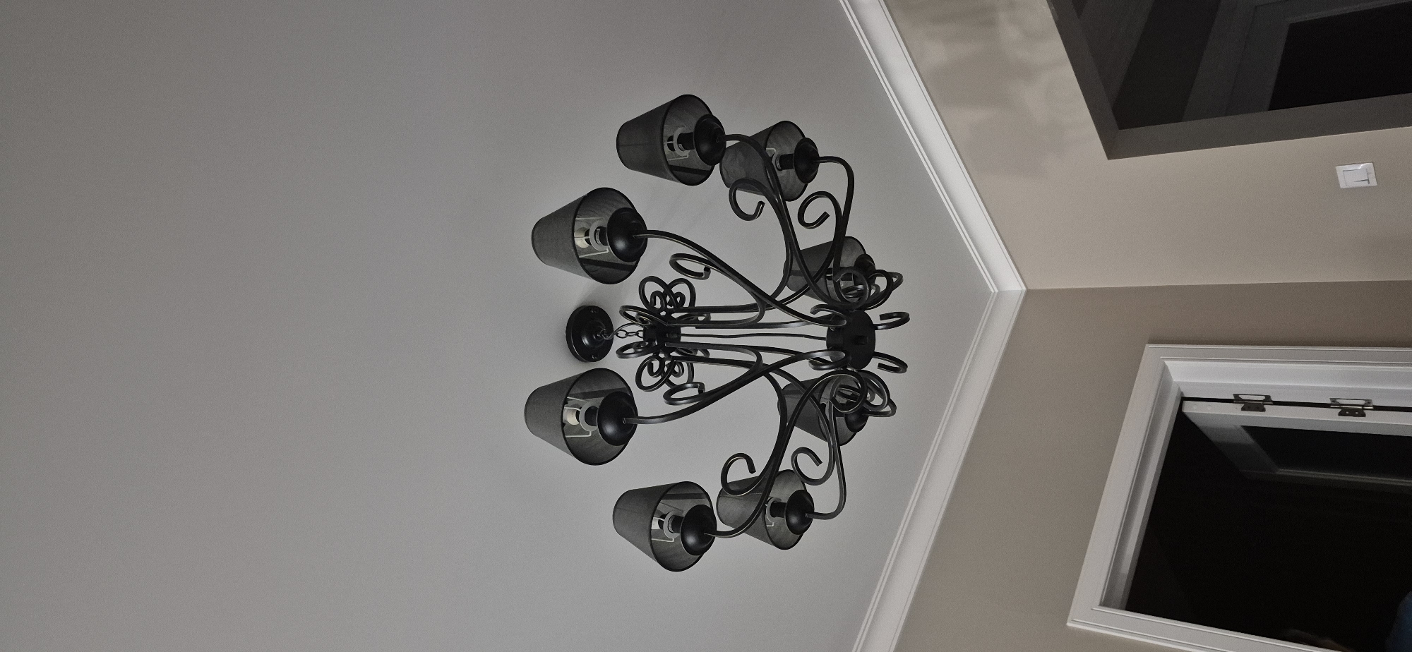 This media file shows an ornate, black wrought-iron chandelier hanging from a white ceiling. The chandelier has multiple arms, each holding a black lampshade with a metallic interior. The overall style of the chandelier suggests it might be a traditional or rustic design.
The setting appears to be indoors, likely a room with a high ceiling, given the perspective and the presence of architectural details like crown molding. The walls are painted a light neutral color, and there's a glimpse of a darker, recessed ceiling or architectural feature in the upper right of the frame. Also visible in the upper right is a white light switch. In the lower right, a portion of a window frame is visible, also in white, with a dark interior suggesting the window is either closed or the space beyond is unlit. The lighting in the scene is dim, casting shadows from the chandelier onto the ceiling, which could indicate it is either nighttime or the room is not brightly lit during the day.
No people are visible in the frame, and there is no discernible text. The focus is on the chandelier and its immediate surroundings, creating a sense of interior design and home decor. The image is taken from a low angle, looking up at the ceiling.