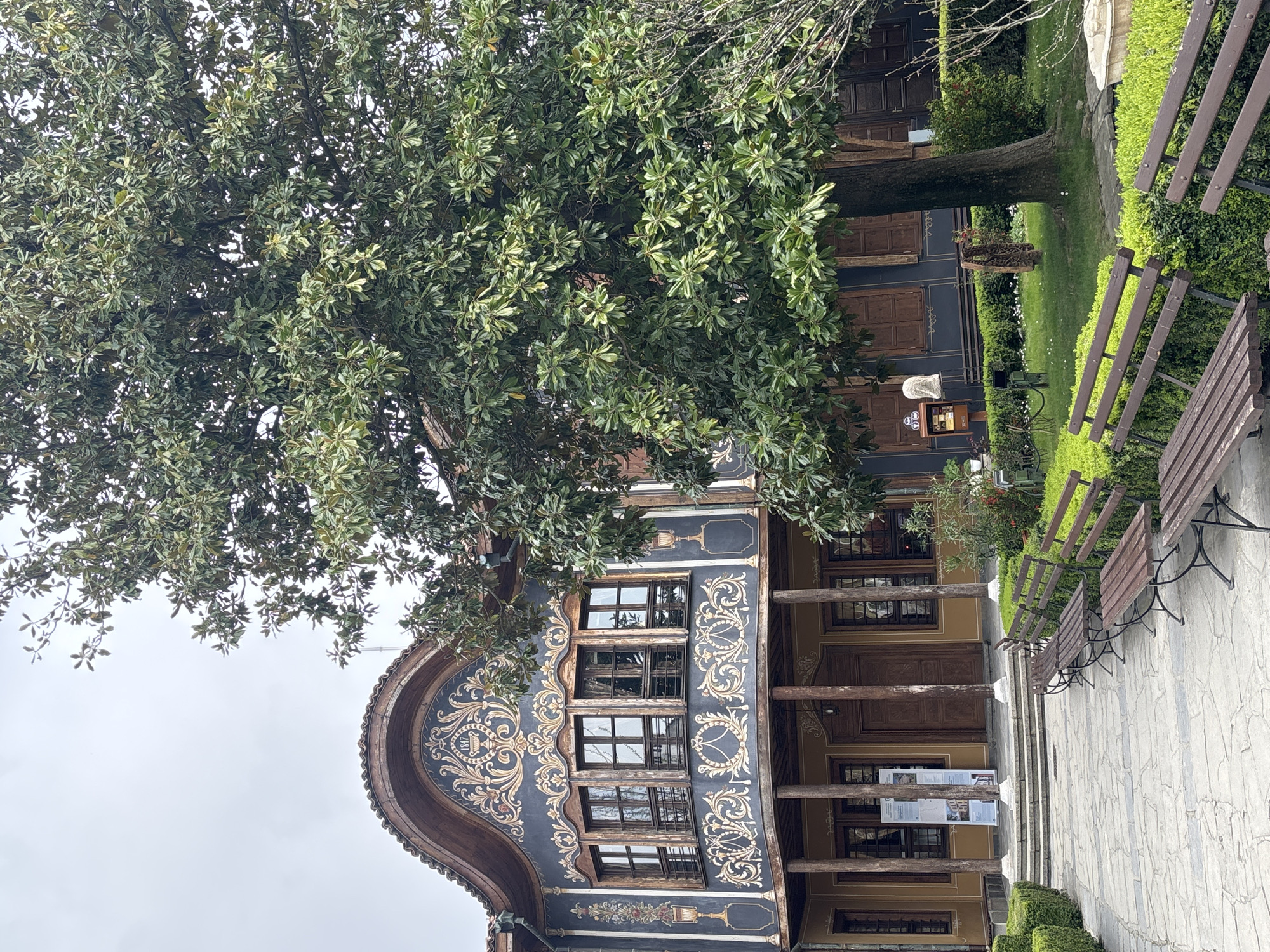 A slightly tilted full shot captures the exterior of a historic building in Plovdiv, Bulgaria, likely a museum or cultural center, partially obscured by a large tree with dense, green foliage. The building features ornate, traditional Bulgarian architecture, characterized by a dark blue facade adorned with decorative, light-colored patterns around the windows. The windows are multi-paned and reflect the cloudy sky. A curved, overhanging structure with similar decorative details tops the building's facade.
Below the windows, a porch or balcony area is supported by wooden pillars. Informational signs are visible on the ground level. To the right of the building, a paved pathway leads away from the camera, flanked by rows of wooden benches and trimmed hedges, suggesting a garden or park-like setting. The sky is overcast, contributing to the muted lighting of the scene. There is no evidence of people, or other animated subjects in the scene.