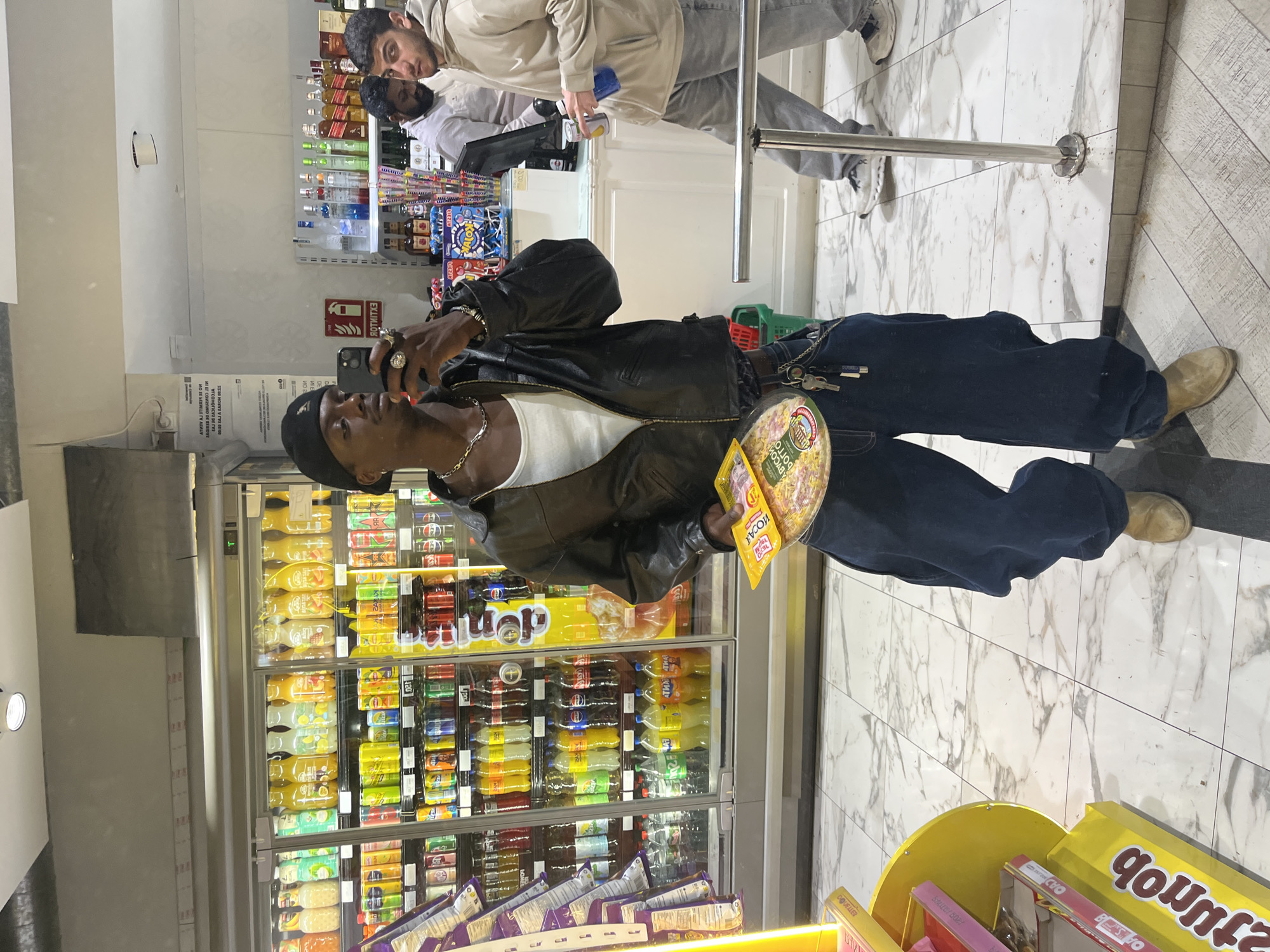 The image depicts a scene inside a brightly lit convenience store or mini-market in Barcelona.
In the foreground, a young Black man is captured in a casual pose. He is wearing a black beanie, a silver chain, a white t-shirt, a black leather jacket, and dark blue, baggy jeans, complemented by light brown boots. He holds a smartphone up, seemingly taking a photo or video, while also clutching a round, flat package, possibly a pizza, with "GOLLO" visible on its label, and a smaller snack packet labeled "KOYALI" and "SMILEY BODY." Multiple rings adorn his fingers, and a chain with keys hangs from his jeans.
In the mid-ground, a large refrigerated display case filled with a variety of beverages like sodas and juices is visible. The floor is covered in white and grey marbled tiles.
Further back, near a checkout counter, two other men are present. One, with a beard, wears a beige hoodie and grey jeans, holding a blue item, while the other, partially obscured, wears a white shirt. Behind them, shelves are stocked with numerous bottles of alcohol. A metal barrier or railing is present near the checkout.
Notable details include a red fire extinguisher sign on the wall, displaying "ROTNITE" (likely "EXTINTOR" backwards). Other signs with partially legible text, potentially in Catalan or Spanish, are visible, such as "ATHIV AJ STIMRI 32 ОИ" which appears to be reversed text. A yellow display stand in the foreground shows "junob" (possibly reversed text for a brand like "BonJour"). The scene captures an everyday moment within a retail setting.