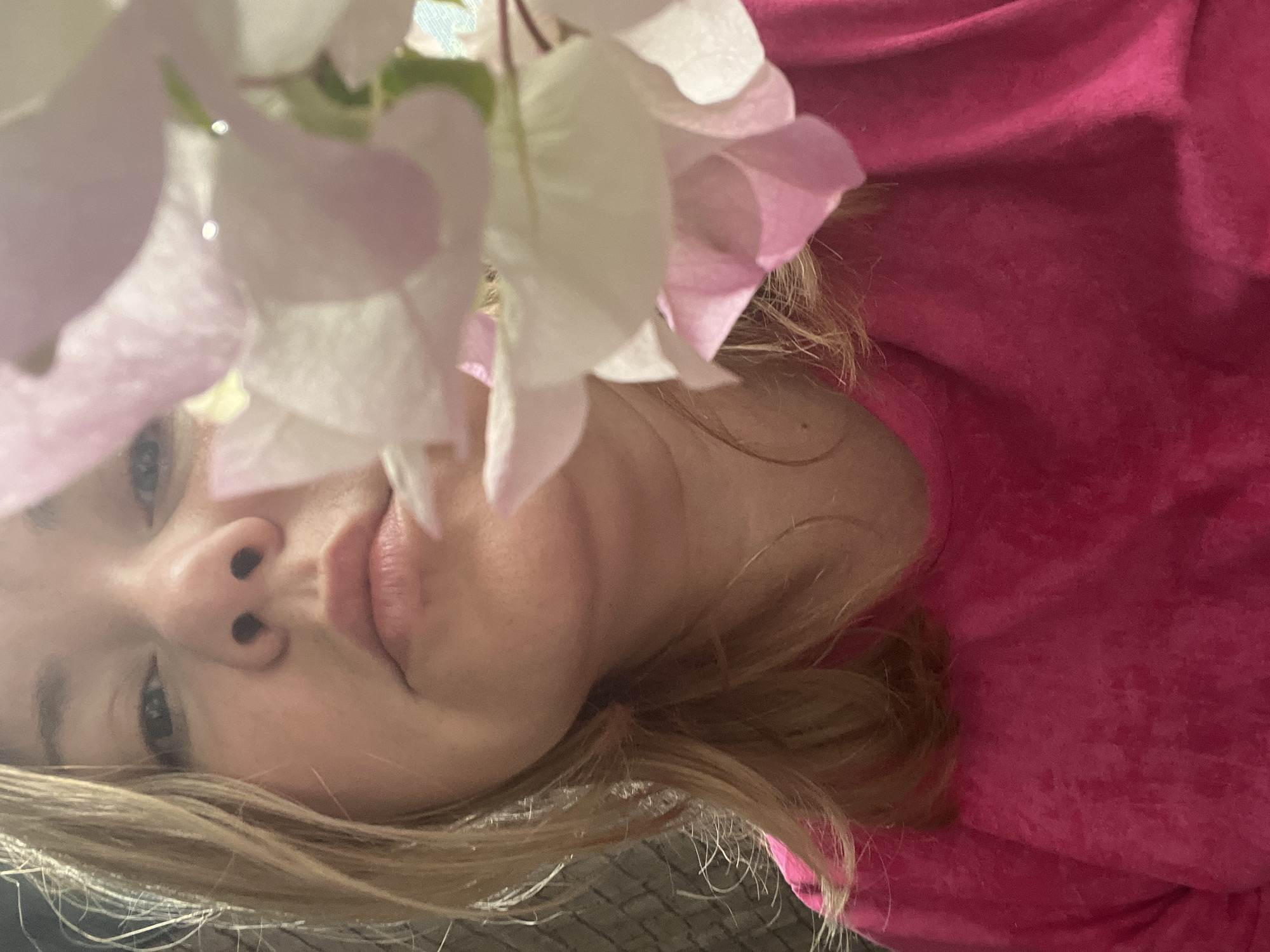 A close-up, slightly angled, indoor portrait captures a woman with light blonde hair. The right portion of the image shows a person in a bright pink garment or clothing. A cluster of white and light pink bougainvillea flowers occupies the top-left and central area of the frame, partially obscuring the woman's face but allowing her eyes, nose, and lips to be visible. The lower-left part of the image shows the woman’s hair over a dark, textured surface. There is no indication of the location of the image or city within the provided information.