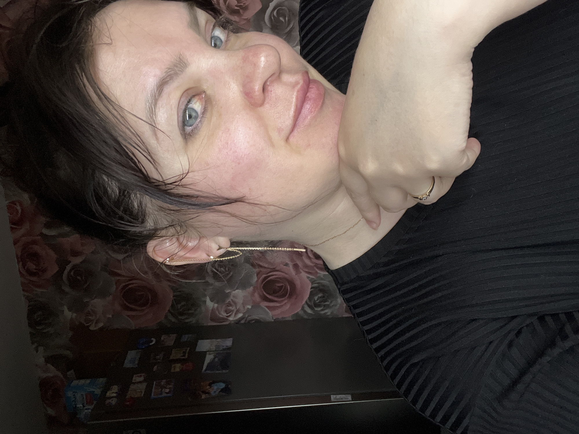 A forensic description of an indoor scene features a woman, appearing to be in her late 20s to early 30s, positioned for a close-up photograph. She has fair skin with localized redness on her cheeks and nose, blue eyes, and dark hair pulled back, appearing damp or styled. Her gaze is directed towards the camera, and her mouth forms a slight, closed smile. Her right hand, adorned with a gold-colored ring on the ring finger, supports her chin. She is wearing a black, vertically ribbed garment and a thin gold-colored chain necklace. A distinctive gold-colored chain earring, spanning from the earlobe to the upper cartilage, is visible on her right ear. The background consists of a wall covered in wallpaper featuring a repeating pattern of large, faded red and pink roses. To the left of the woman, a dark, vertical surface, possibly a domestic appliance or cabinet, is partially visible, bearing multiple small, rectangular items resembling photographs or magnets, and a light blue and white package near the top. The precise location within Noviy, Russia, is not discernible from the visual evidence presented in the image.