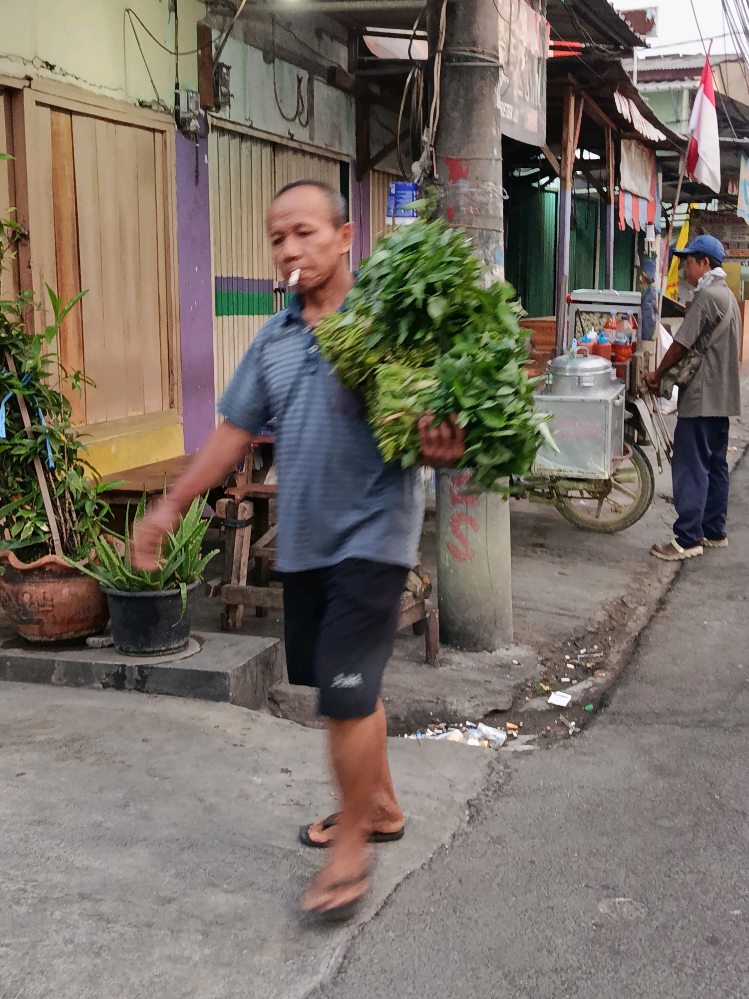 A man with a cigarette in his mouth walks carrying a large bundle of green leafy vegetables. He is wearing a striped blue polo shirt and black shorts. He is walking on a paved sidewalk. In the background, there is a street vendor with a cart, and a building with a corrugated metal roof. The scene appears to be a street market in Bekasi, Indonesia.