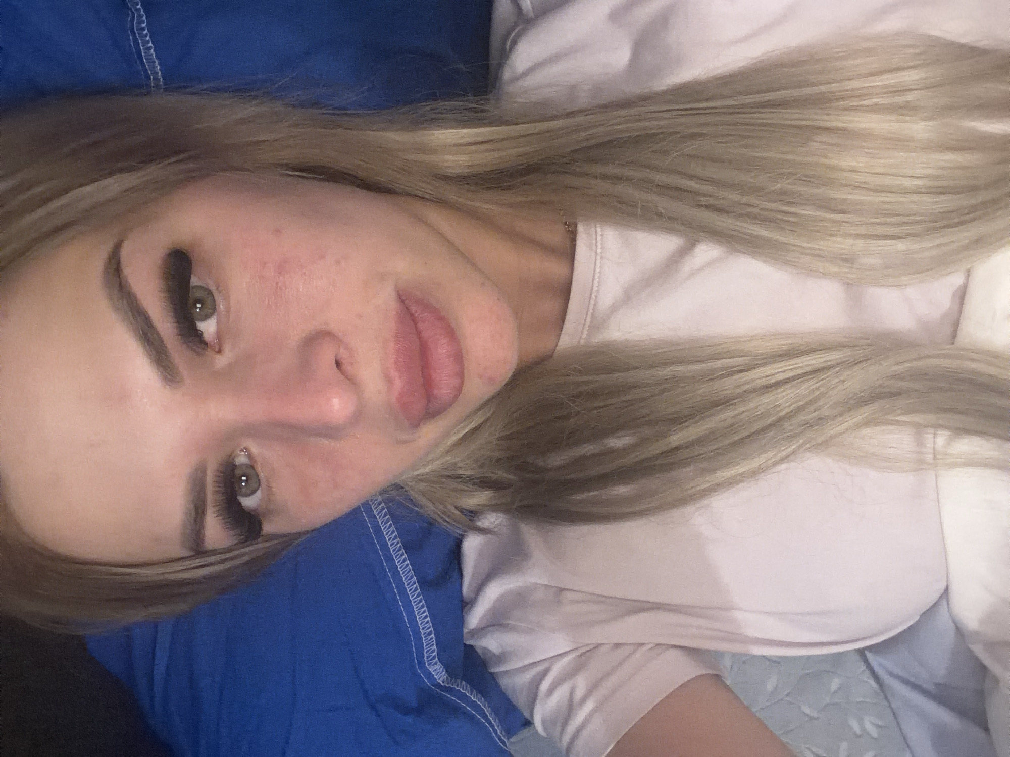 A close-up, overhead shot shows a fair-skinned woman with blonde hair, lying down, possibly in a bed. Her face is angled slightly to the right, with her eyes open and looking at the camera. She is wearing a light pink t-shirt. Her hair is long and straight, falling down around her shoulders. A bright blue pillow is visible behind her head. The background also includes white bedding. The image appears to be taken indoors, and there are no discernible landmarks or environmental cues to suggest a specific location in Kyiv, Ukraine, or any other city.