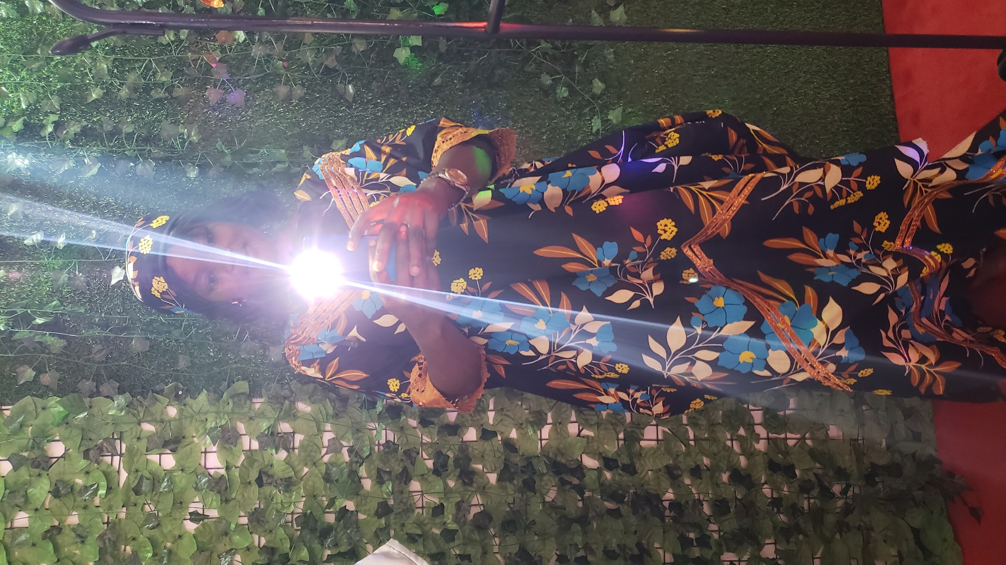 A dark-skinned woman stands in front of a backdrop of artificial green ivy and grass. She is wearing a black dress with a floral pattern of blue flowers and yellow accents. She also has a matching headscarf. The woman is holding a smartphone in both hands, and the screen is illuminated, casting a bright light on her face and the front of her dress. A vertical black pole, possibly part of a photography setup, is visible to the right of the woman. The floor is covered in red carpet. This image was taken in the city of Kinkinau, Nigeria.