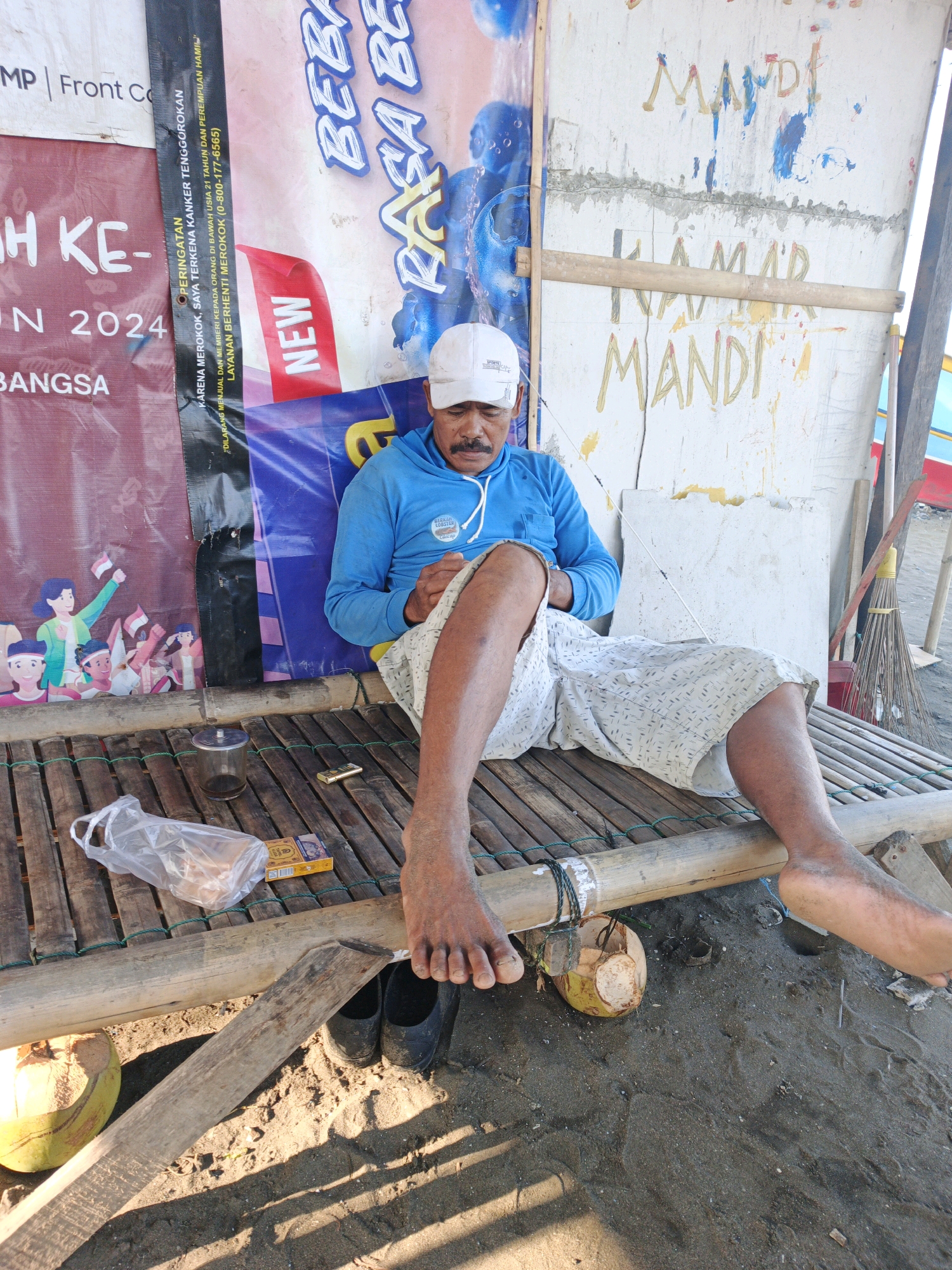 The image depicts a man sitting on a bamboo platform on a sandy beach. The man is wearing a blue hoodie and white shorts, with a white baseball cap on his head. He has a mustache and appears to be looking down at his hands. In front of him on the platform are a glass of liquid, a pack of cigarettes, and a plastic bag containing some items. To his right, a broom leans against a white wall with some colorful markings and writing. In the background, banners with text are visible. A coconut is placed on the sand to the left of the platform, and a pair of black shoes are on the sand beneath the platform. The scene appears to be outdoors, likely in a coastal area of Kelurahan Cilacap, Kecamatan Cilacap, Indonesia, given the textual context on the banners and the beach environment. The overall atmosphere is relaxed and informal.