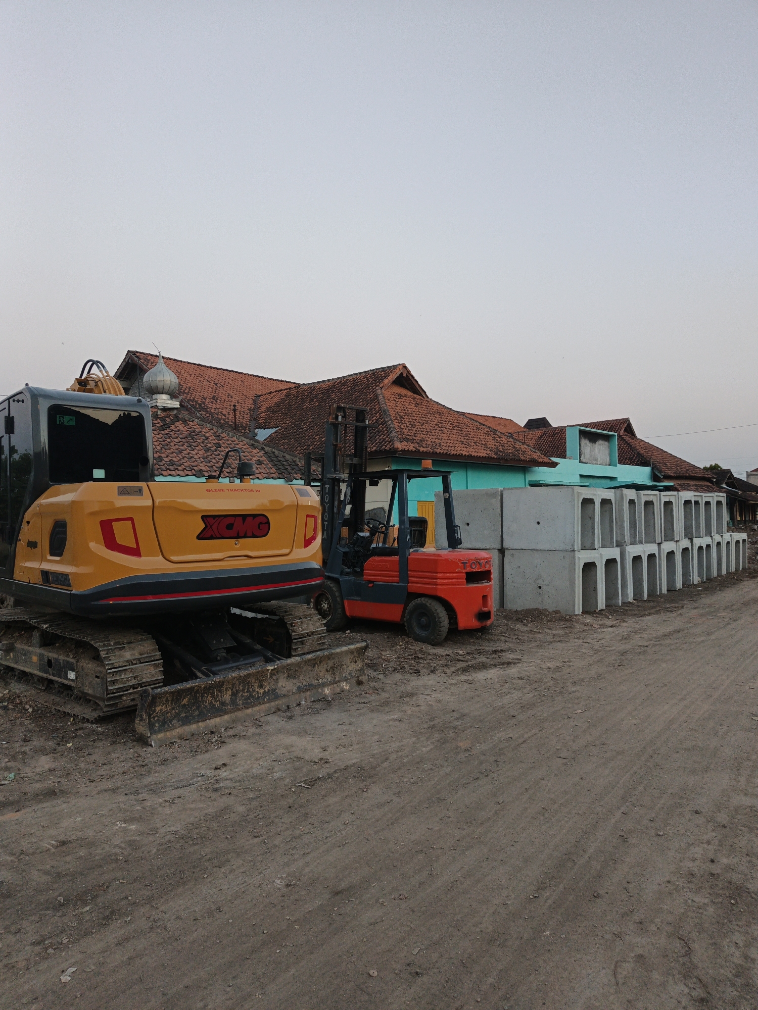 In Planggu, Indonesia, a construction site is depicted under a pale sky. On the left, a yellow and black XCMG excavator is parked on a dirt surface, its tracks showing signs of use. Adjacent to it, a red and black Toyota forklift stands ready. In the background, a row of precast concrete drainage channels is stacked, with a long, low building featuring red-tiled roofs and turquoise walls behind them. The ground is uneven and dusty, suggesting an active or recently used construction area.