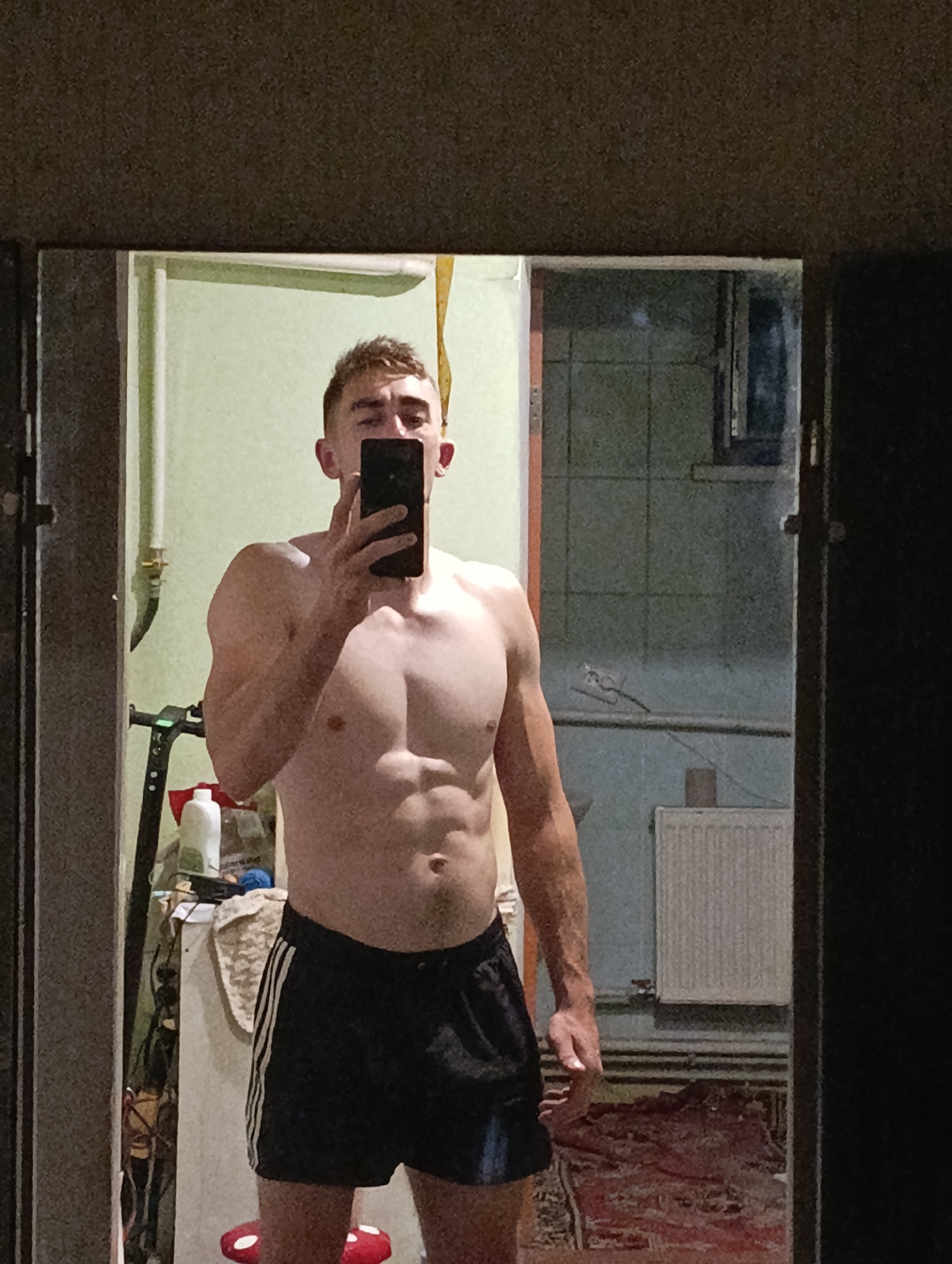 A shirtless person with muscular definition, wearing black athletic shorts with white stripes, stands in front of a mirror, holding a black smartphone to take a selfie. The person's reflection is visible, showing a portion of their face, torso, and legs. The background in the reflection appears to be an indoor setting, possibly a bathroom or bedroom, with a wall-mounted radiator, some shelves with bottles, and a folded rug on the floor. The overall lighting is dim. The location is identified as Fastiv, Ukraine.