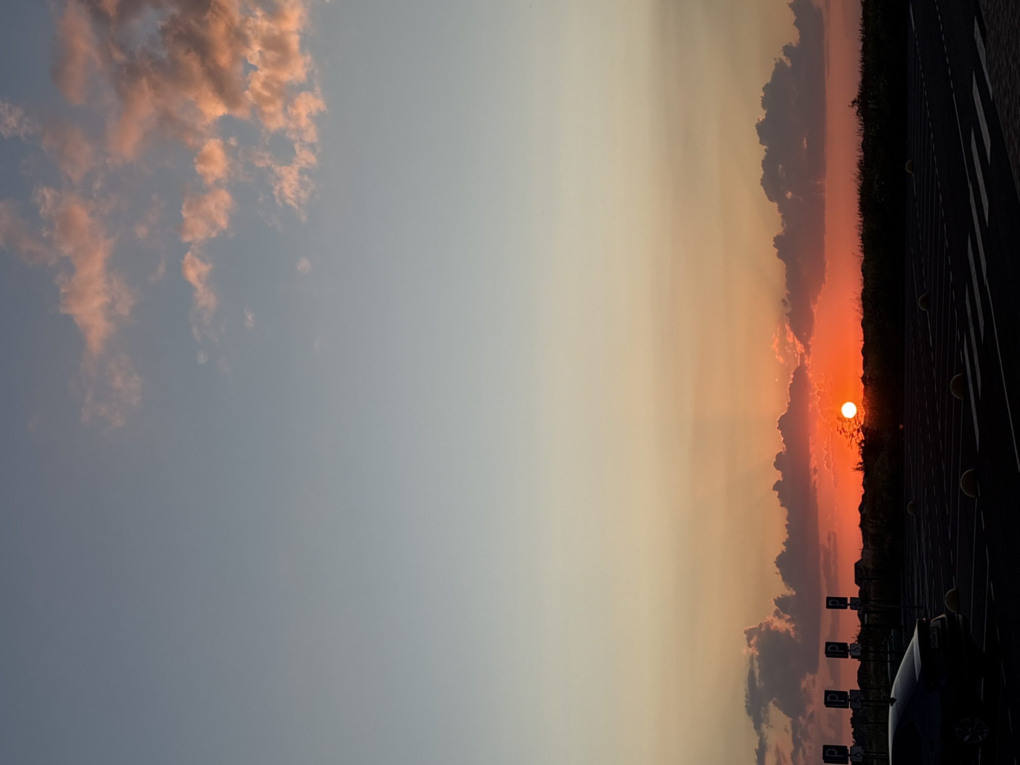 This image depicts a sunset over a parking lot with a line of parked cars and road markings. The sun, a bright orange orb, is visible on the horizon, partially obscured by clouds. The sky transitions from a pale blue on the left to vibrant orange and red hues near the sun. Some clouds are tinged with pink and orange, illuminated by the setting sun. In the foreground, a dark-colored car is parked on the right side of the frame, with other cars and parking signs visible further down the lot. The ground is paved, with clear white markings indicating parking spaces. A dark, silhouetted line of trees and possibly buildings forms the distant horizon. The overall scene evokes a sense of calm and the beauty of a natural sunset. The time of day is clearly evening, as indicated by the sunset. There is no visible text in the image.
