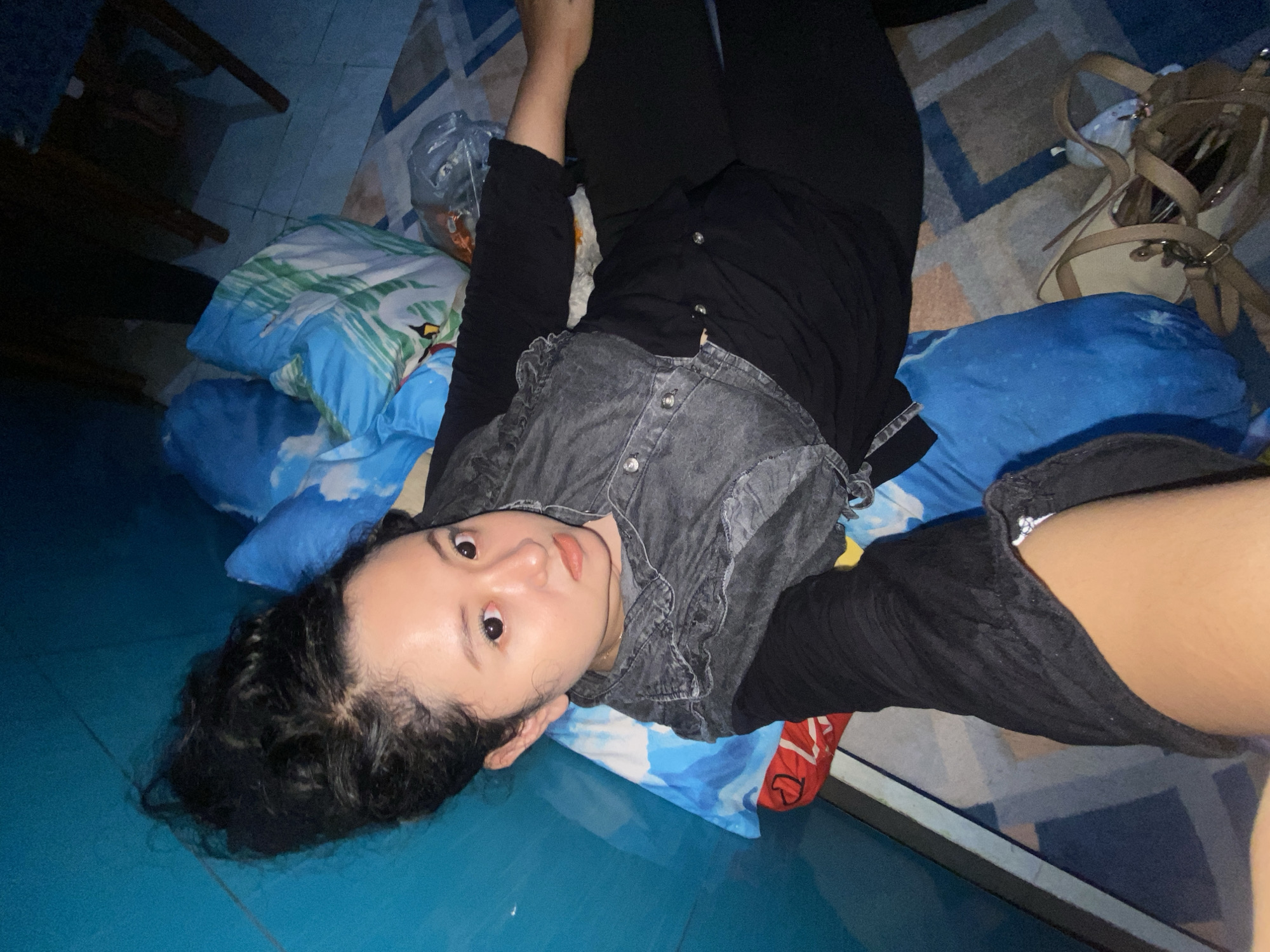 A young woman lies on her back, partially propped on blue patterned pillows. She wears black pants, a long-sleeved black shirt, and a gray denim vest. She looks directly at the camera. Her hair is black and pulled back. She lies on a blue tiled floor, which extends into the background. In the upper right corner, a tan purse with multiple straps sits on a patterned rug. A plastic bag filled with items is visible to the left of her head. A chair leg is visible in the upper left corner. This scene is in an unknown location in Gelam, Indonesia.