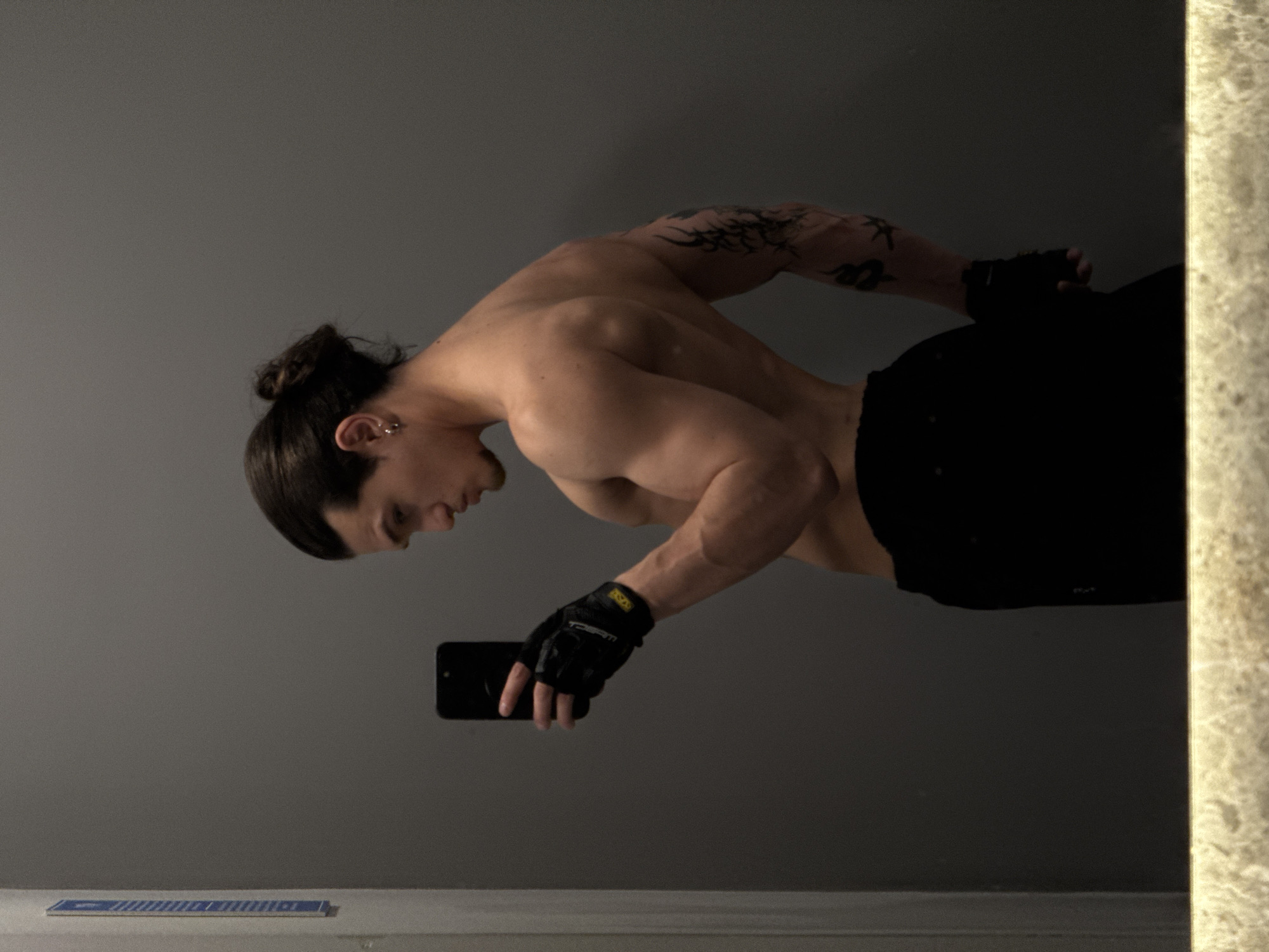 The image, presumably a mirror selfie, depicts a person with a muscular physique in an indoor setting. The individual, appearing male, has long dark hair styled in a bun and wears small stud earrings on their left ear. Visible tattoos adorn their left arm, featuring a tribal-style design on the shoulder/bicep and a snake-like motif on the forearm. They are wearing black fingerless gloves, likely for weightlifting, with yellow accents and partially visible text, and black shorts or pants. The person holds a black smartphone in their right hand, aiming it towards the mirror.
The primary activity is taking a self-portrait, with the subject striking a pose that accentuates their well-defined back and arm muscles. They are seen bending slightly forward, with their head turned towards their right hand holding the phone, looking downwards.
The background is a plain, dark gray wall. To the right, a vertical strip of light-colored, textured material, possibly marble or stone, frames the scene, indicative of a mirror's edge or an adjacent architectural feature. In the bottom left, a thin, rectangular object with blue and white stripes is placed on a light-colored surface, possibly a counter or shelf.
Notable details include the subject's prominent musculature, particularly the sculpted shoulders, trapezius, and deltoids. Soft, even lighting illuminates the scene, highlighting the contours of the body. The text "TECHM" is discernible in yellow on the left glove. The individual's expression is neutral, conveying focus on the photography. The time of day is not discernible due to the indoor lighting. The location context, Sofia, Bulgaria, is provided but not visibly indicated within the image itself.