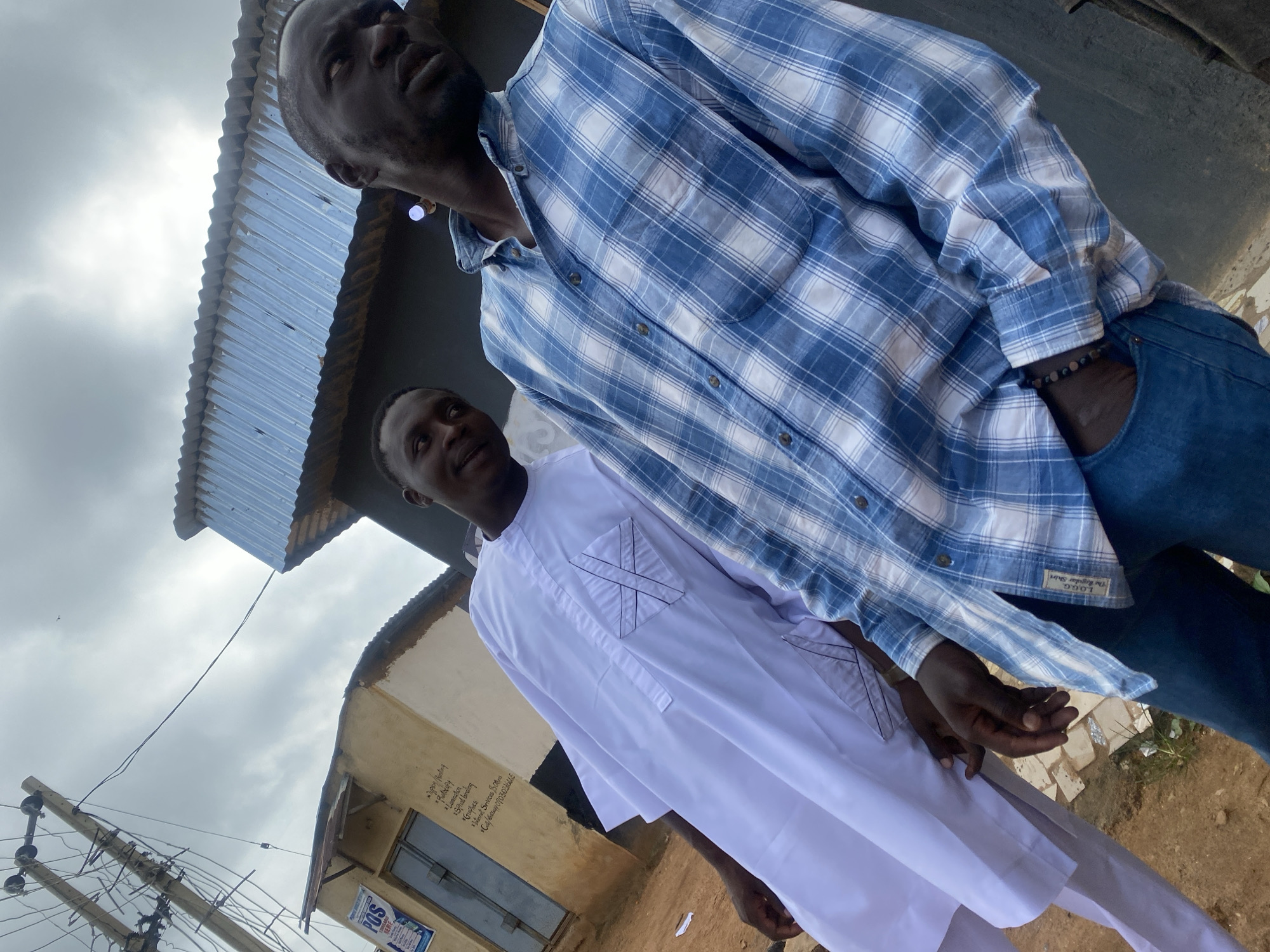 Two men are standing in front of a small commercial building with a corrugated metal roof. The man on the left is wearing a white traditional African garment with black embroidery on the chest pocket. The man on the right is wearing a blue and white plaid button-down shirt and blue jeans. He has a beaded bracelet on his left wrist. The building has signs on its exterior, one of which reads "POS" and lists services like "Digital Printing," "Photocopy," "Lamination," "Spiral binding," "Graphics," "Internet Services," and a phone number. The environment appears to be an outdoor, urban setting, possibly in the city of Rafin Par, Nigeria, characterized by a cloudy sky, a utility pole with wires, and a dirt and gravel ground.