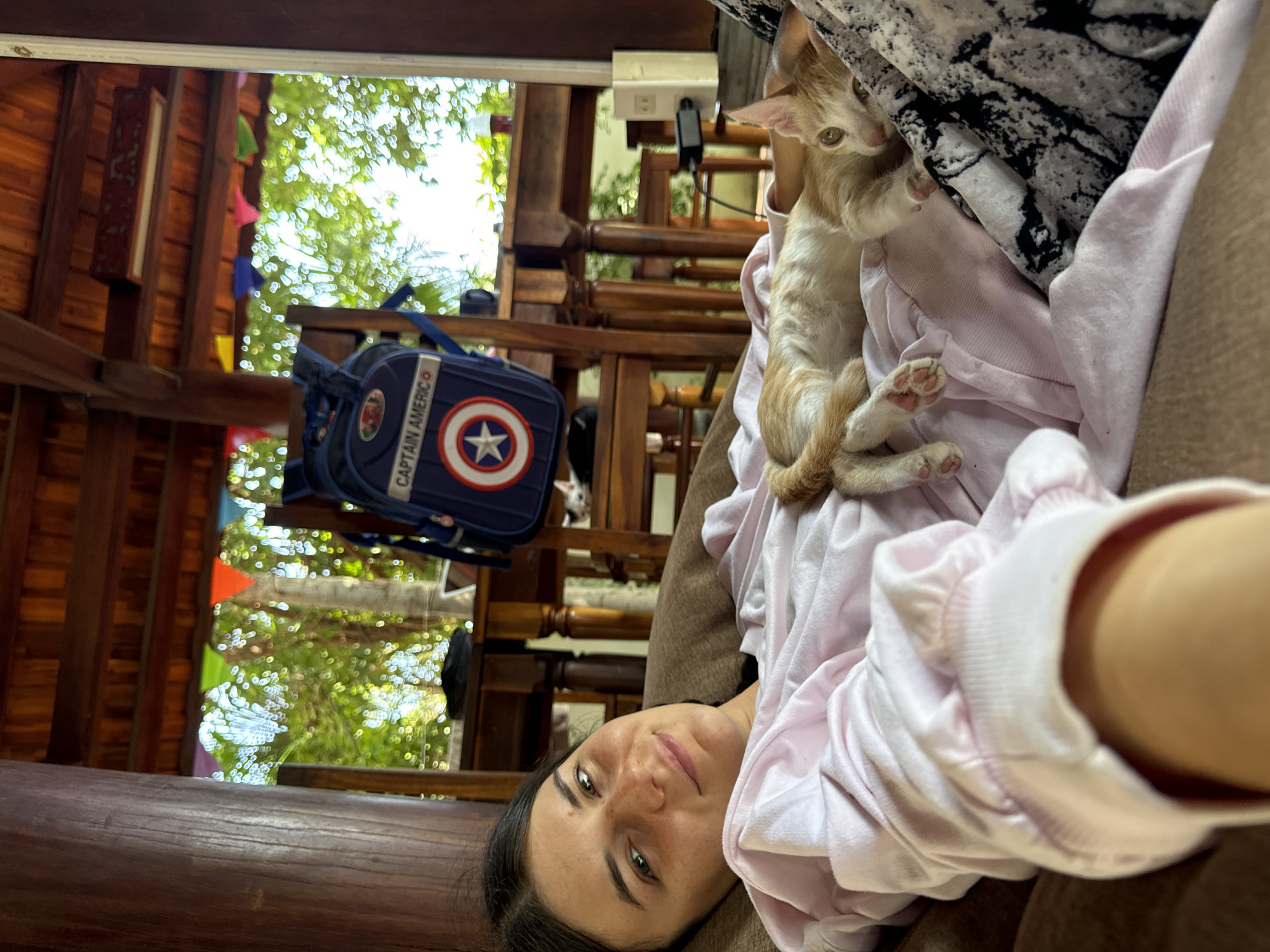 A young woman with dark hair and light skin lies on a brown couch in an open-air structure. She is wearing a light pink sweatshirt. Next to her, a ginger and white kitten sleeps on her pink sweatshirt. In the background, a blue backpack with a Captain America emblem is propped against a wooden chair. Beyond the chairs, lush green foliage is visible through an opening in the structure, suggesting an outdoor location. The ceiling is made of dark wooden beams. The setting appears to be a casual, relaxed environment, possibly a resort or home in Mũi Né, Vietnam, given the tropical vegetation and open architecture.