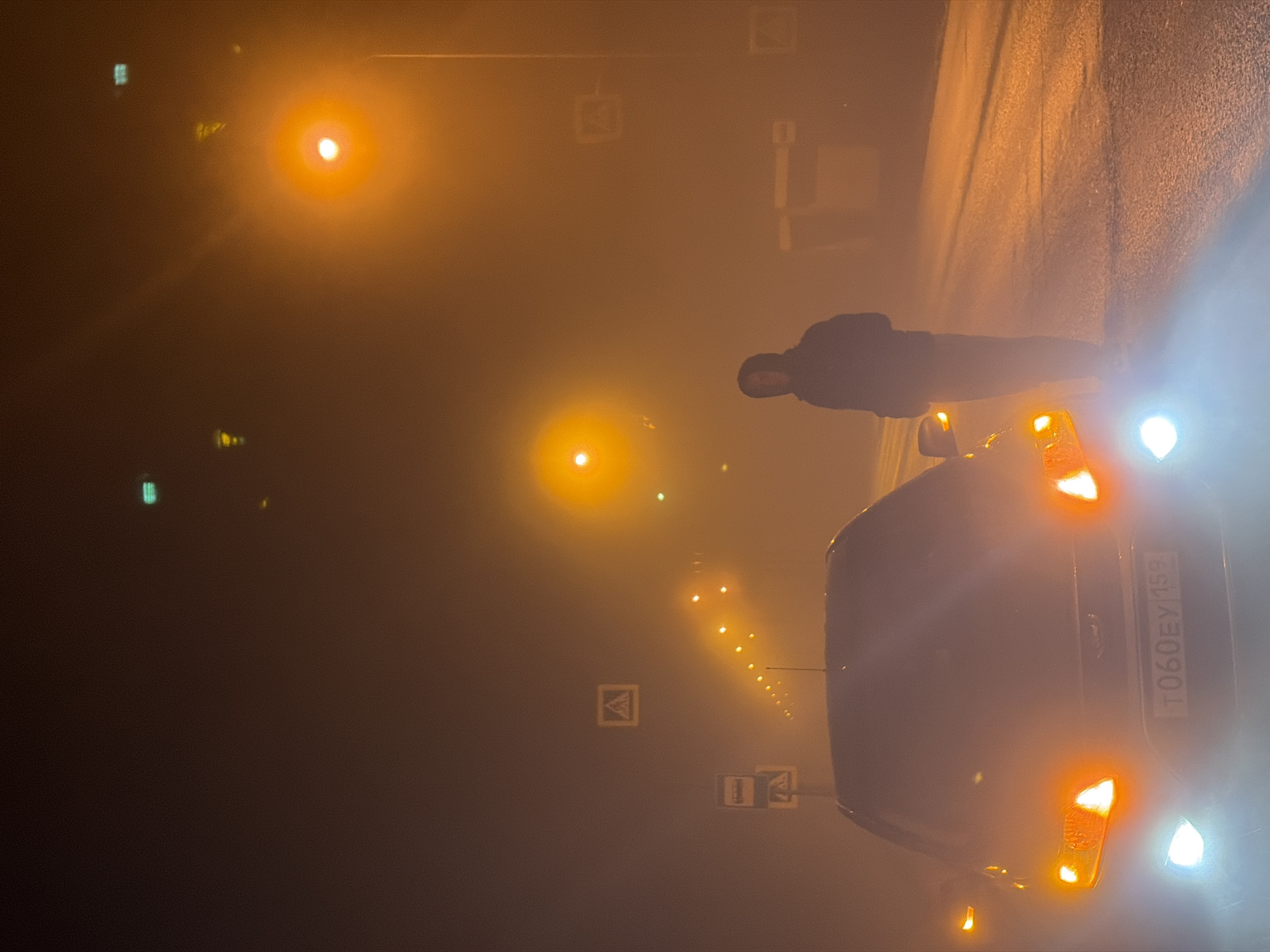 A nighttime image shows a person next to a car on a road in Noviy Urengoy, Russia. The road is wet and reflects light. The car's license plate is visible with the number "T060EY159." The scene is foggy, with streetlights illuminating the road in an orange glow. Several road signs are visible, although obscured by the fog. The person stands beside the car, their form obscured by the poor visibility. The car's headlights and blinkers are illuminated.