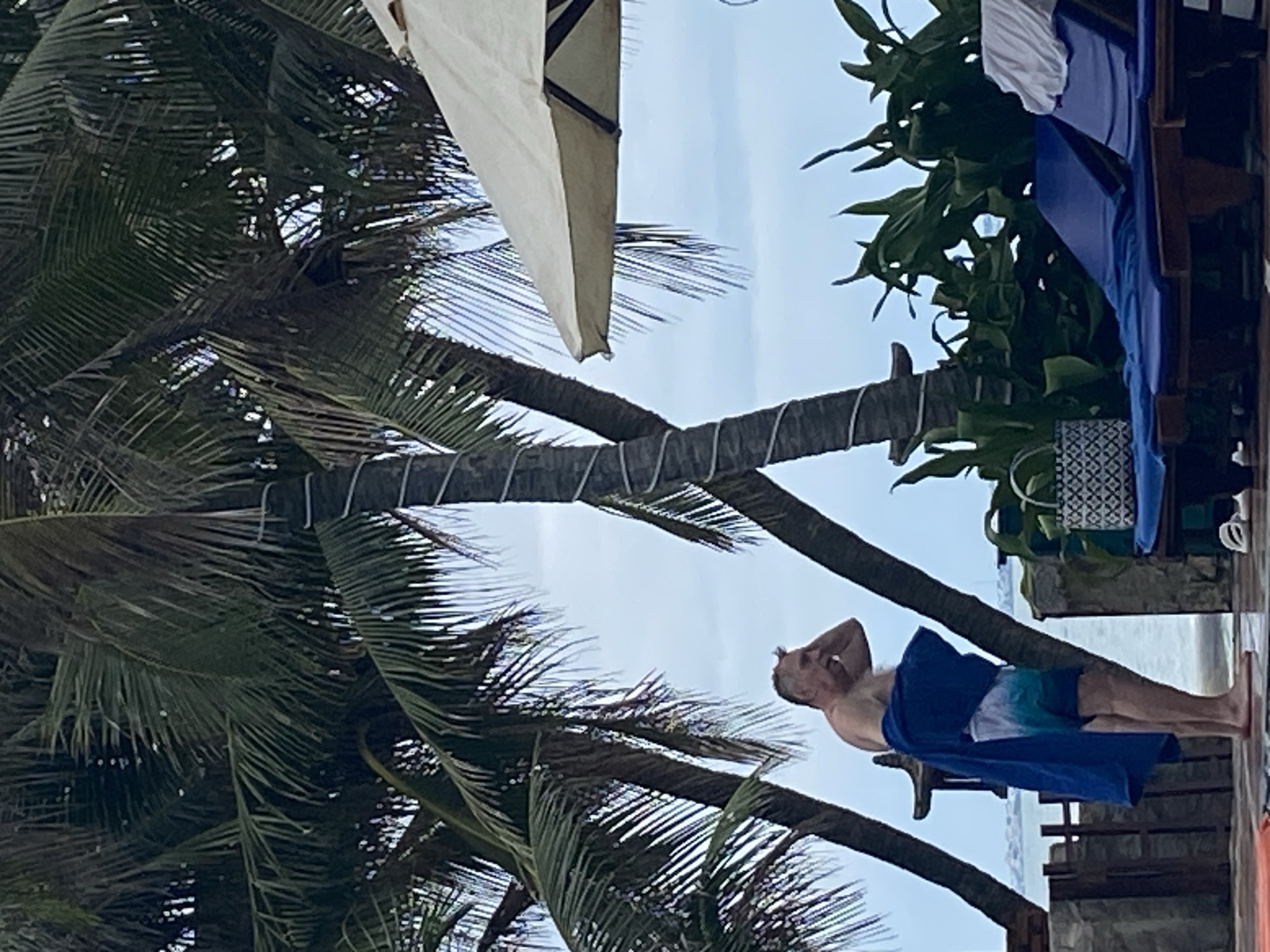 A beach scene in Phu Thuy Ward, Vietnam features a man standing near a wooden structure with a blue towel draped around him, his hand raised to his head. The backdrop is filled with palm trees, some secured with rope, and an open-air umbrella. The sky is visible in the gaps between the trees.