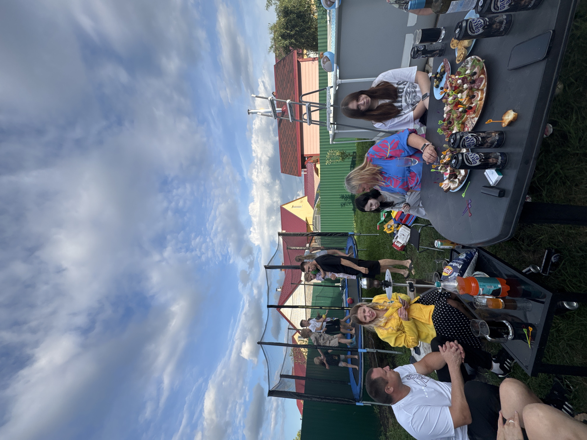 In a backyard gathering in Uzuyukovo, Russia, several people are enjoying an outdoor party. A large trampoline is set up with multiple children actively jumping. Around a low table, adults are seated and engaged in conversation, with drinks and food laid out. The table features cans of "Green Bear" beer, a bottle of orange soda, and plates of appetizers. Nearby, a portable swimming pool with a ladder stands, and the surrounding environment includes a fence, houses with red roofs, and a cloudy sky. The scene appears to be a casual social event.