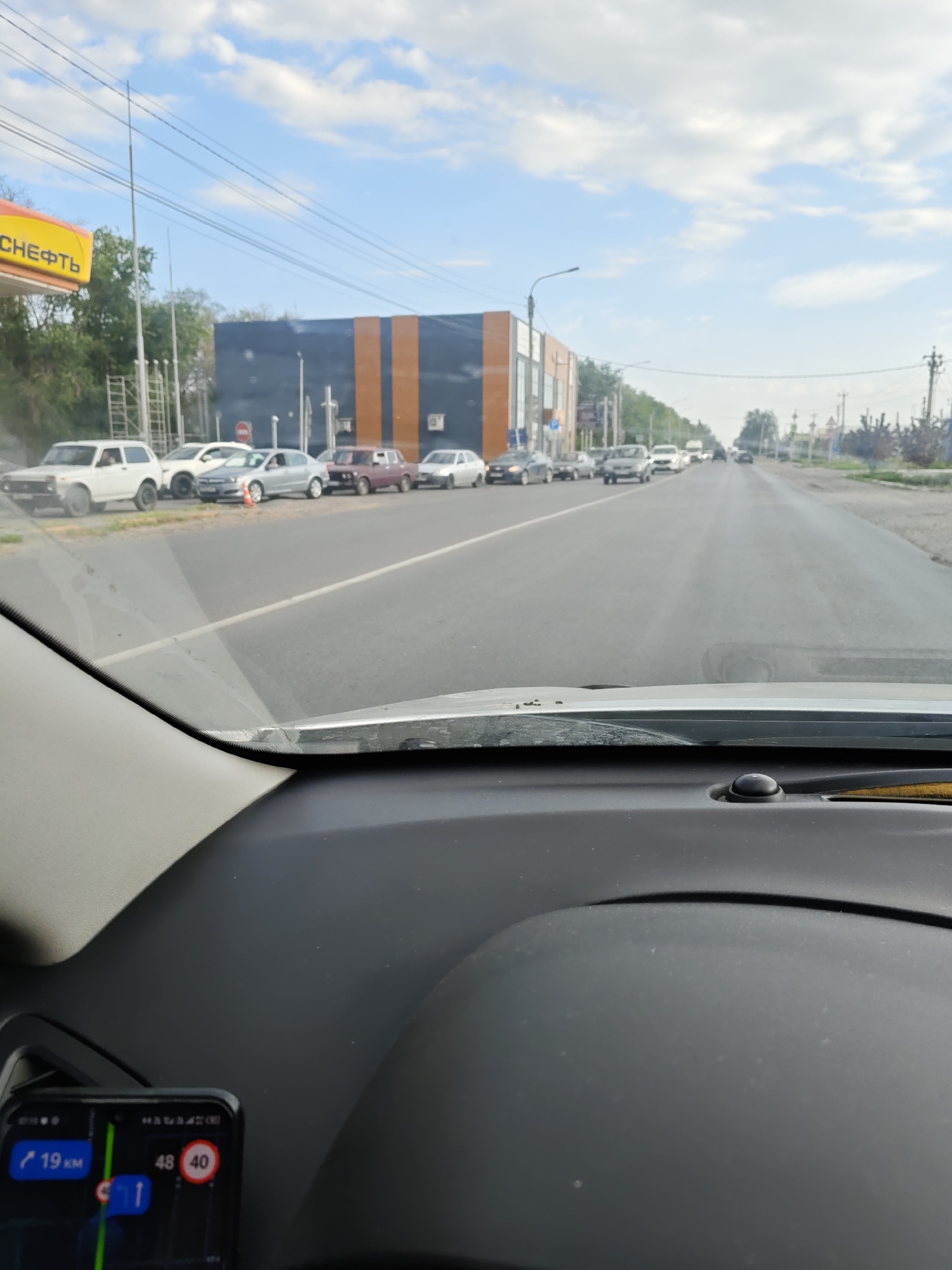 This is a first-person view from inside a vehicle, looking out at a street scene in Novocherkassk, Russia. A yellow sign with "НЕФТЬ" (oil) written on it is visible on the left. The road ahead is lined with parked cars and other vehicles in traffic. A modern building with dark blue and orange sections is prominent on the left side of the street. The sky is partly cloudy. A smartphone mounted on the dashboard displays a navigation app with speed limit information (40 km/h).