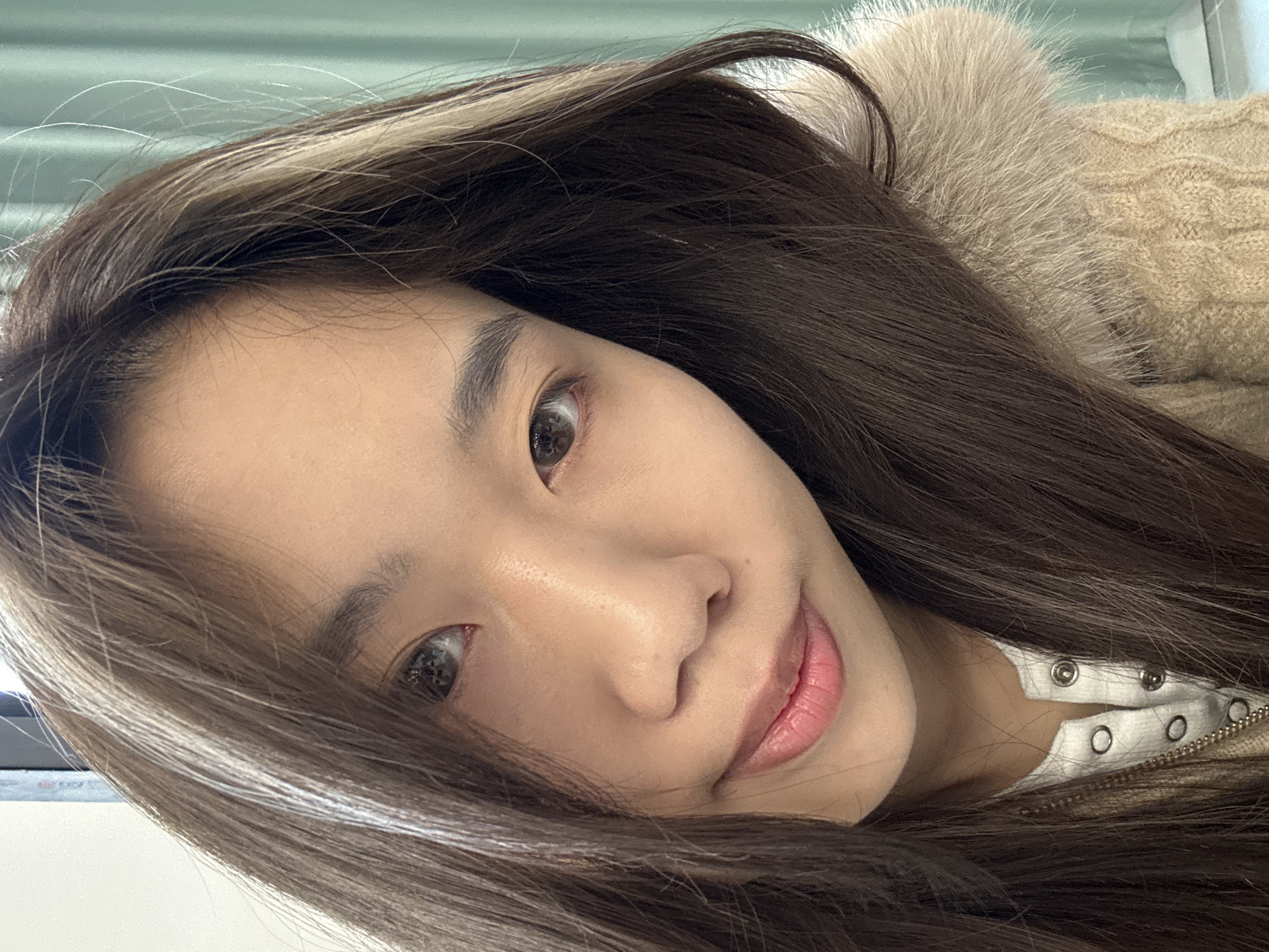 A close-up portrait depicts an East Asian female from the upper chest upward. She has long, dark brown hair with lighter, possibly highlighted, streaks flowing over her shoulders. Her eyes are dark brown, directed at the viewer, and her lips form a subtle, closed-mouth smile. She wears a light-colored, possibly white or light grey, top with silver snap buttons visible on the placket, beneath a beige cable-knit garment featuring a light-colored fur-like collar on the right side of the frame. A silver chain or zipper pull is partially visible on her left chest. The background is an out-of-focus indoor environment, presenting a white wall or panel on the left and a light green or teal curtain or blind on the right. The image contains no identifiable features to determine a specific location within Nan'an District, China.