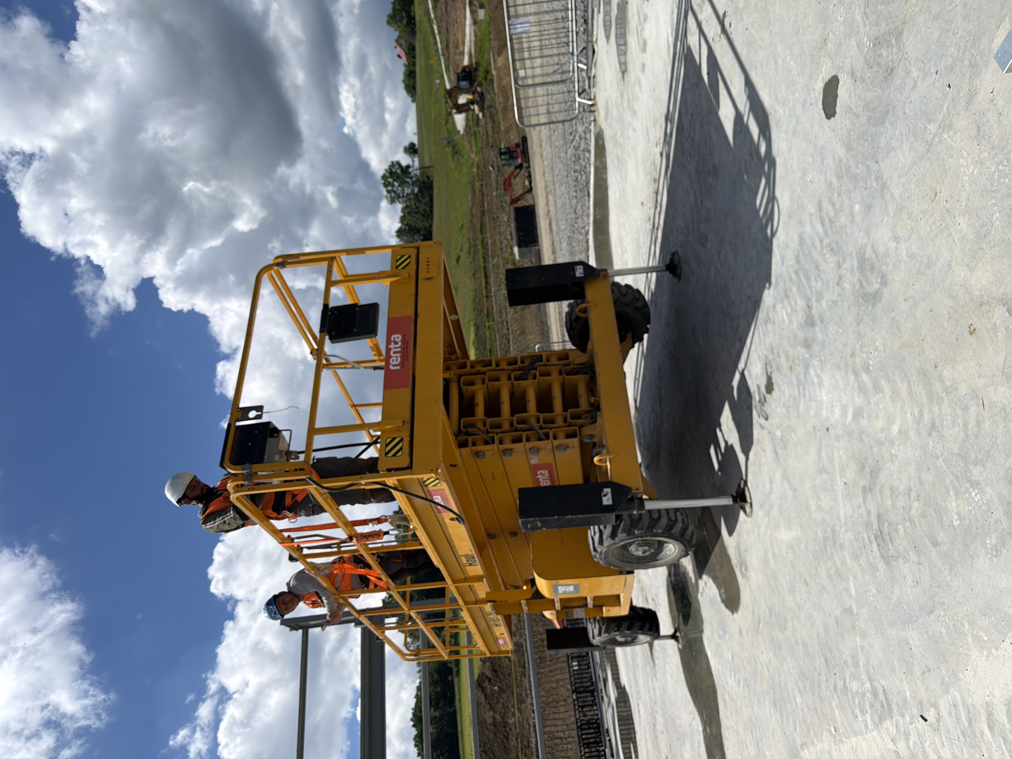 Two construction workers, wearing hard hats and high-visibility vests, stand on the platform of a yellow scissor lift at a construction site. One worker is looking down at a device, possibly a tablet or control panel, while the other is looking towards the camera. The scissor lift is positioned on a concrete surface, with its stabilizers deployed. In the background, there is a hillside with trees and some buildings. The sky is blue with scattered white clouds. Several excavators and construction barriers are visible in the middle ground, suggesting ongoing construction activity. The scene appears to be a sunny day, as indicated by the shadows cast by the scissor lift and its occupants. The location is identified as Janowice, Poland.
