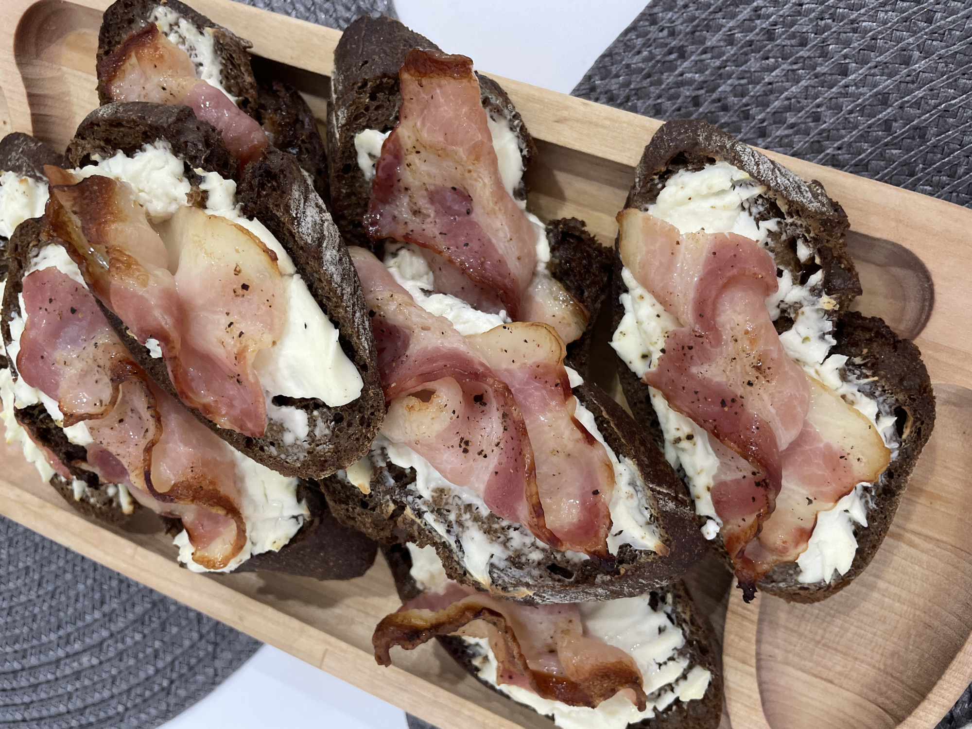 The image displays a wooden tray holding several slices of dark bread topped with a creamy white spread and strips of cooked bacon. The bacon pieces are scattered and layered on each slice, with black pepper visible on some of them. The tray rests on a surface that is partly white and partly covered with a gray woven mat. The scene appears to be a casual food presentation or serving arrangement. The image does not contain any information related to the city of Izhevsk, Russia.