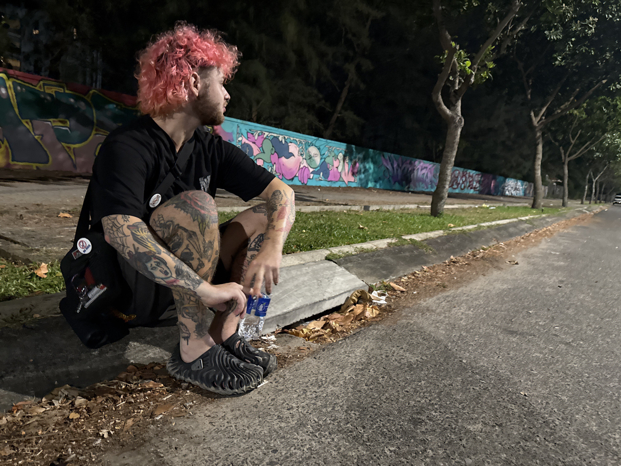 A man with pink hair sits on a curb next to a street in Đà Nẵng, Vietnam. The man is wearing a black t-shirt and shorts and has extensive tattoos on his arms and legs. He is holding a water bottle. To his left is a black bag adorned with buttons. Behind him is a wall covered in colorful graffiti. A row of trees lines the street, and a car can be seen in the distance. The scene appears to be at night.