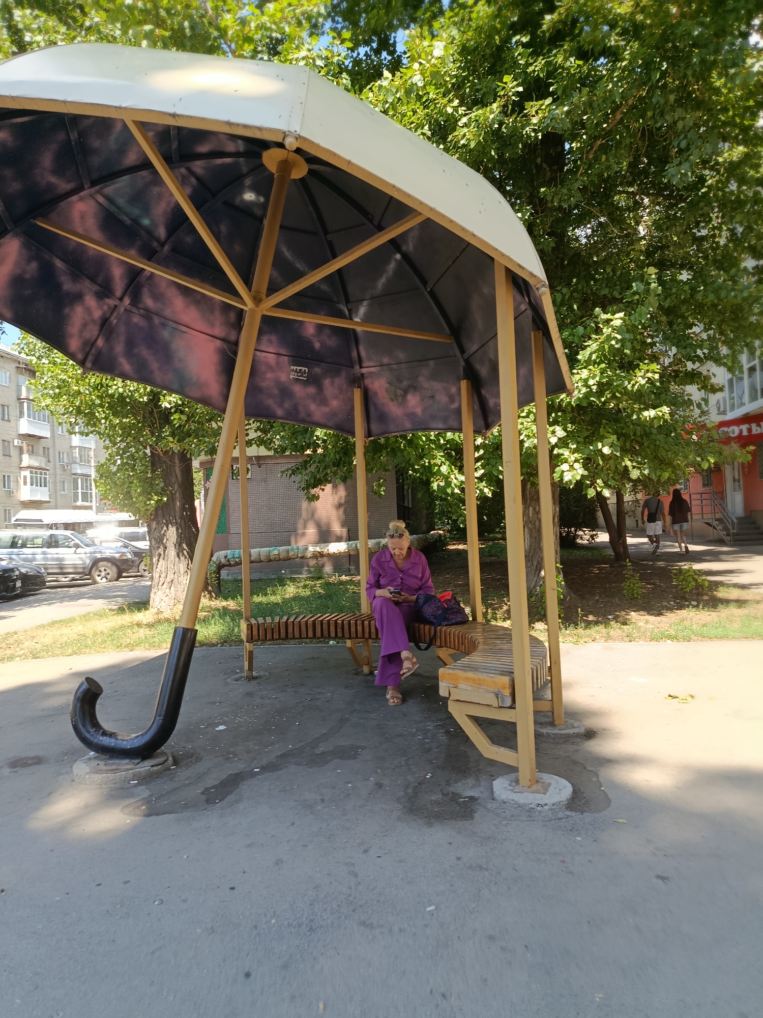 A woman, likely of retirement age, is sitting on a wooden bench inside a large, umbrella-shaped shelter. She is wearing a purple jumpsuit and sandals and is looking at her phone. The shelter has a dark canopy and yellow poles. A large, curved black handle resembling that of an umbrella is visible on the ground to the left of the shelter. The ground is paved with asphalt. In the background, trees with green foliage line a street, and a multi-story building is visible. Cars are parked along the street. The scene appears to be a public space, possibly a bus stop or a rest area, in the city of Saratov, Russia. The overall impression is one of a sunny day in an urban environment.