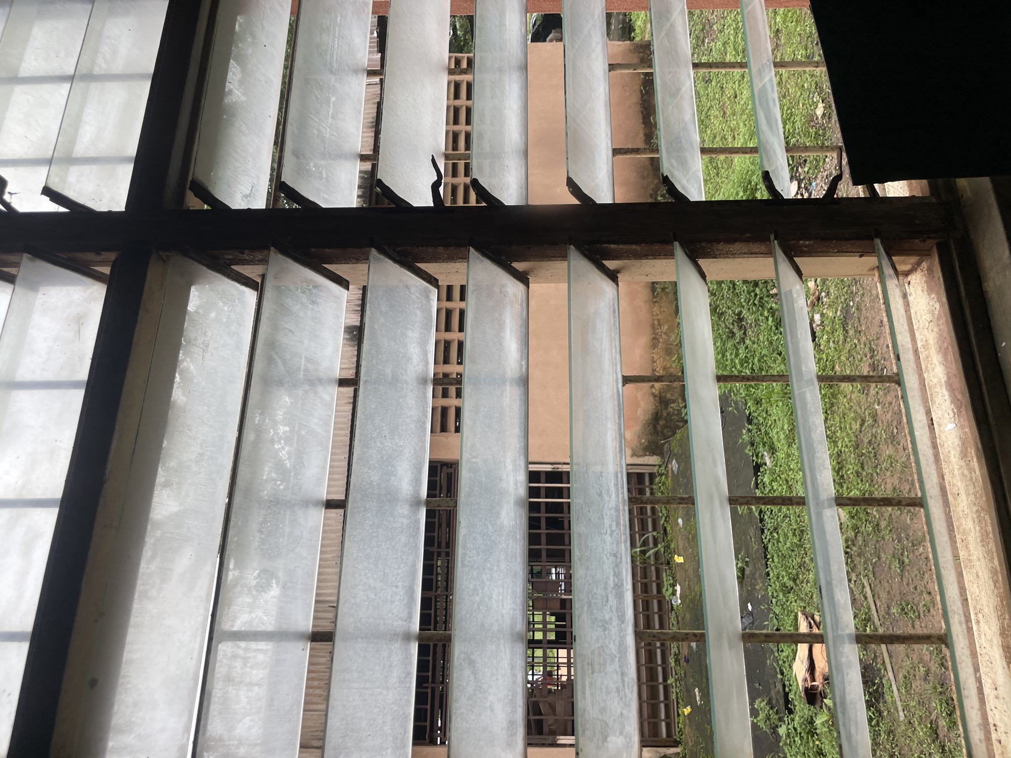 This image shows a louvre window with glass slats, viewed from inside a building in Ihiagwa, Nigeria. The window overlooks an outdoor area with overgrown green vegetation, scattered debris, and a concrete path. Behind the vegetation, a building with orange-colored walls and barred windows is visible. The louvre window itself has wooden frames and metal supports holding the glass slats at an angle, allowing for ventilation. The lighting suggests it is daytime, possibly overcast, as the light is diffused. The scene depicts an exterior view through a functional, possibly aging, window.