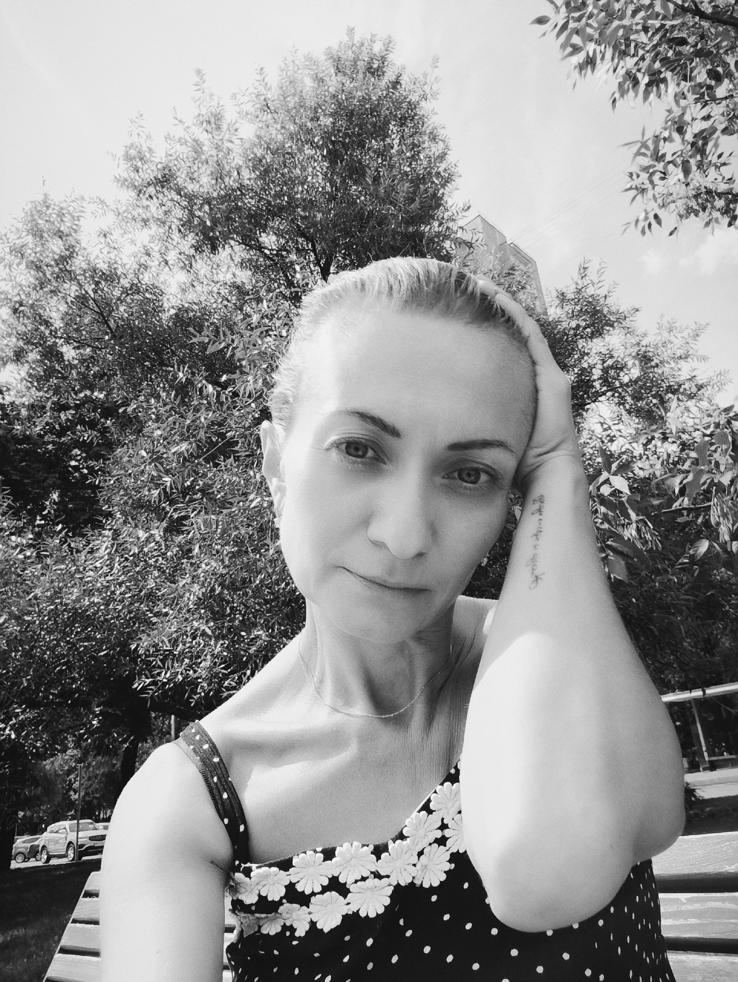 A black and white, medium shot, selfie captures a woman outdoors. She is positioned in the foreground, leaning her head on her right arm, which is raised and bent at the elbow. Her left arm is resting on her lap or on the bench she is seated on. She has blonde hair pulled back, and her gaze is directed towards the camera. A delicate necklace is visible around her neck. Her attire consists of a sleeveless dress with a polka dot pattern and floral embellishments around the neckline. A tattoo is visible on her right forearm.
The background features lush green foliage from trees, suggesting a park or a natural setting within a city. In the distance, a building structure can be partially seen, and a few cars are parked. The lighting indicates it is daytime, with the sun casting shadows. The image appears to have been taken in a city park, likely in Moscow, Russia, given the commonality of such park settings in the city.