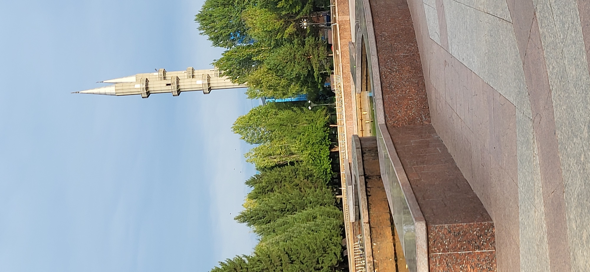 The image depicts an outdoor scene in the city of Oskemen, Kazakhstan, focusing on a multi-tiered fountain and a minaret. The fountain features several levels with granite-like cladding, and the water within appears calm. Behind the fountain, a line of trees, a mix of coniferous and deciduous, forms a green backdrop. In the distance, a tall, slender minaret, characteristic of mosque architecture, rises against a pale blue sky with scattered clouds. The minaret is light-colored, possibly concrete or stone, and has multiple balconies or viewing platforms. The ground in the foreground is paved with large, square tiles in varying shades of gray and brown. The overall impression is one of a public space, likely a park or square, designed for relaxation and contemplation, with the minaret serving as a prominent landmark.