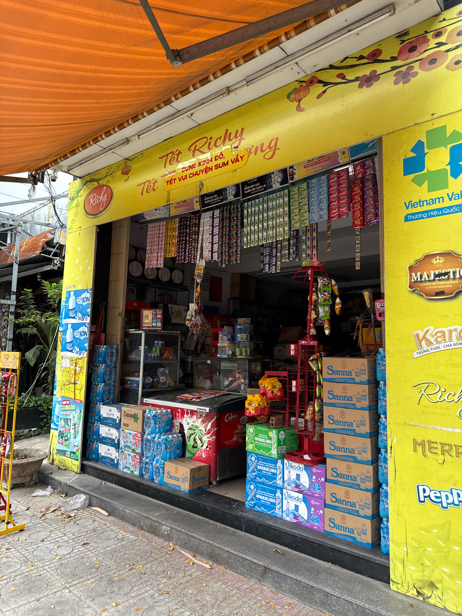 This image shows a street-level convenience store in Nha Trang, Vietnam, likely decorated for the Tet holiday, as indicated by "Tet" prominently displayed on the yellow awning. The store has an open facade, with products displayed both inside and outside. On the left, a yellow support column is covered with posters, and a yellow display rack holds bagged snacks. Inside the store, various drinks are stacked, including cases of Aquafina water and Sanna water bottles. Shelves are stocked with packaged goods, and a red display rack in the center holds smaller snack bags. A red and white refrigerator is visible at the back right of the store. The store exterior is painted bright yellow with red and floral decorations. The ground in front of the store is paved with gray tiles and is littered with fallen leaves and a few pieces of plastic trash. The overall impression is that of a small, busy neighborhood shop.