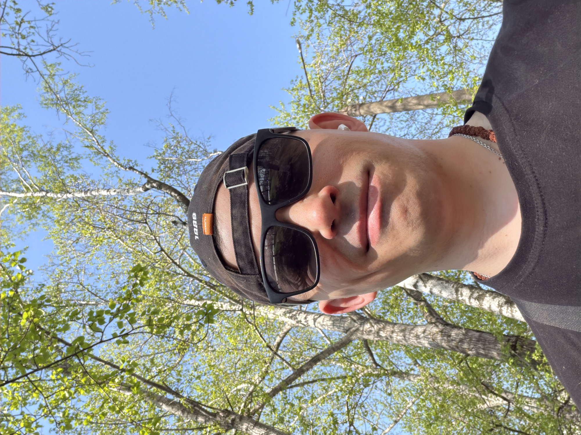 The image is a selfie of a person wearing a black baseball cap and sunglasses, positioned looking up at the sky and tree branches. The person is wearing a black shirt. The setting appears to be outdoors, with green leaves and tree branches visible against a blue sky. The location in Izhevsk, Russia, cannot be determined based solely on the information in the image.