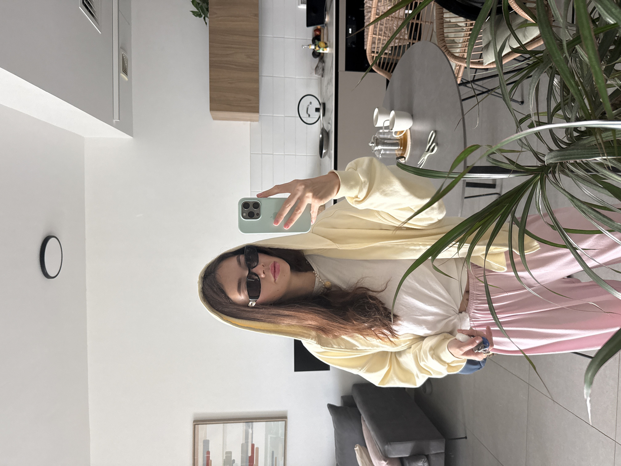 A woman is taking a selfie with a light blue smartphone. She is wearing sunglasses, a yellow hoodie, a white t-shirt, and pink sweatpants. The location appears to be an interior space, with a kitchen visible in the background. A round table and a wicker chair are situated near a plant. A sofa and a framed picture are visible in the background, suggesting a living room area. The city and specific location within the city are unknown.