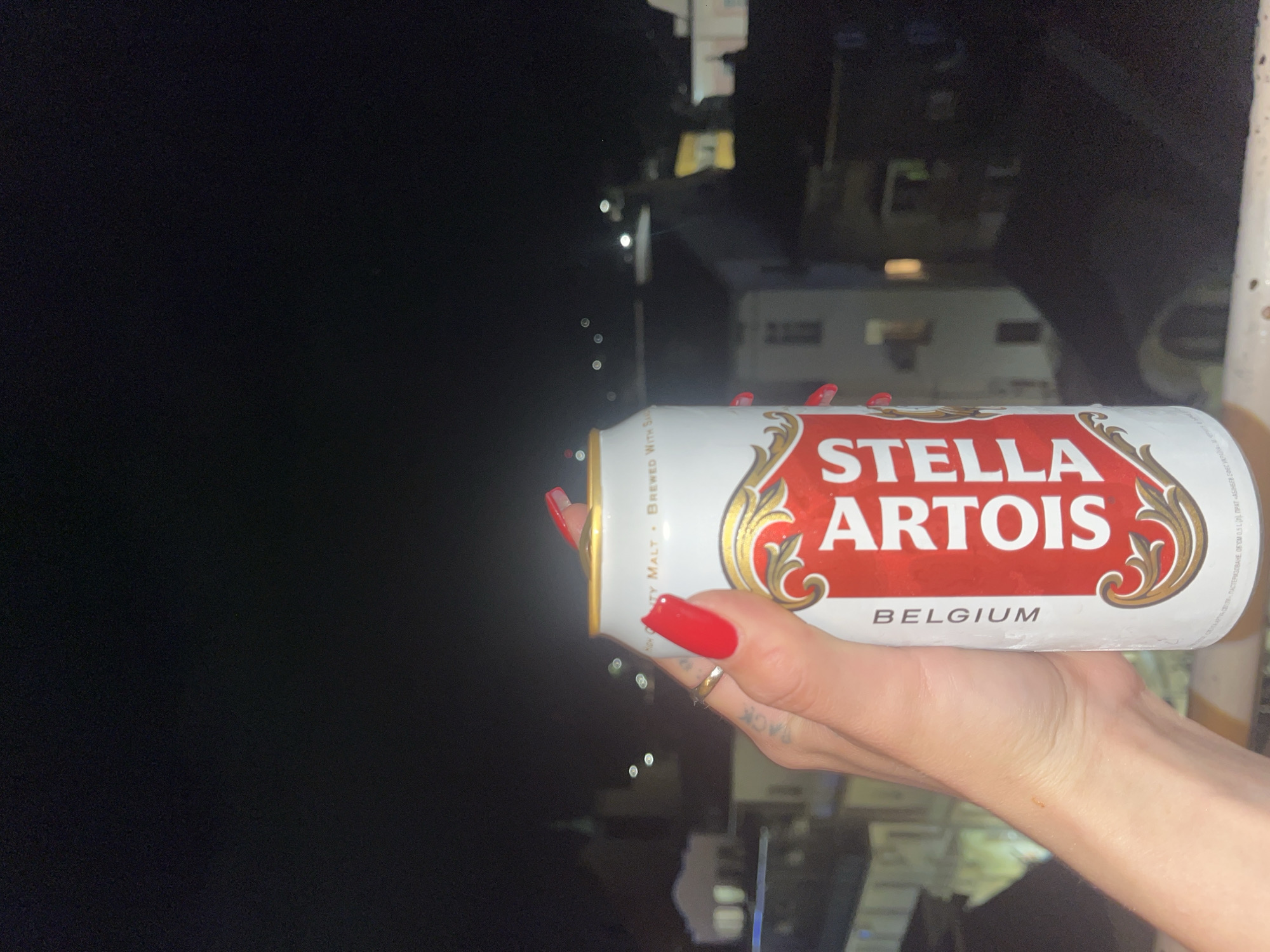 The image displays a close-up of a person's right hand holding a Stella Artois beer can, oriented horizontally. The hand has long, bright red fingernails and wears a gold-colored ring on the ring finger. A tattoo, partially visible as "BACK," is on the side of the index finger. The Stella Artois can is predominantly white with a distinctive red and gold label, featuring the brand name "STELLA ARTOIS" and "BELGIUM." Additional text on the can includes "NTY MALT," "BREWED WITH SA," and Ukrainian script indicating "PASTEURIZED," "VOLUME 0.5 L," and the producer "AB INBEV EFES UKRAINE."
The setting is outdoors at night, characterized by a dark, out-of-focus background. Numerous small, blurry lights are scattered across the left side of the frame, with indistinct, darker architectural shapes visible on the right. This suggests an urban or semi-urban environment, possibly viewed from an elevated position such as a balcony. The activity is simply holding the beverage. No specific emotions are discernible, but the scene conveys a casual, relaxed atmosphere in the evening.
