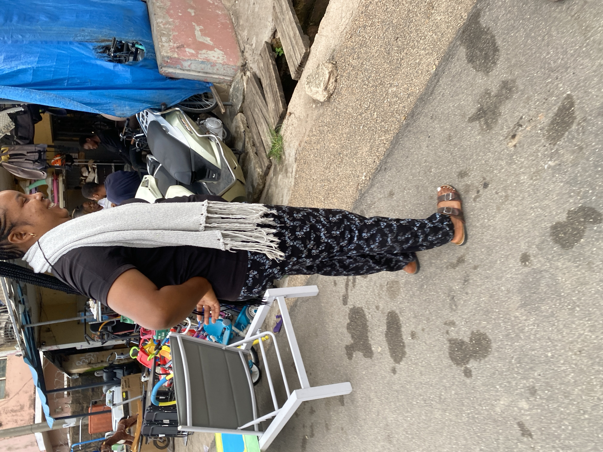 The image shows a woman standing on a street in Jos, Nigeria. She is wearing a black t-shirt, patterned pants, a grey scarf, and brown sandals. Her hair is in braids. She appears to be looking to her left, with a slight smile.
Behind her, there is a market stall with various items, including what appears to be a scooter, children's toys, and chairs. A blue tarp is draped over some of the goods. The ground is asphalt with some wet patches. In the background, there are buildings and what looks like a drainage ditch with wooden planks.
The scene suggests a bustling market environment. The lighting and shadows indicate it is daytime, possibly late morning or early afternoon. The overall impression is that of an everyday scene in a Nigerian marketplace. There is no visible text in the image.